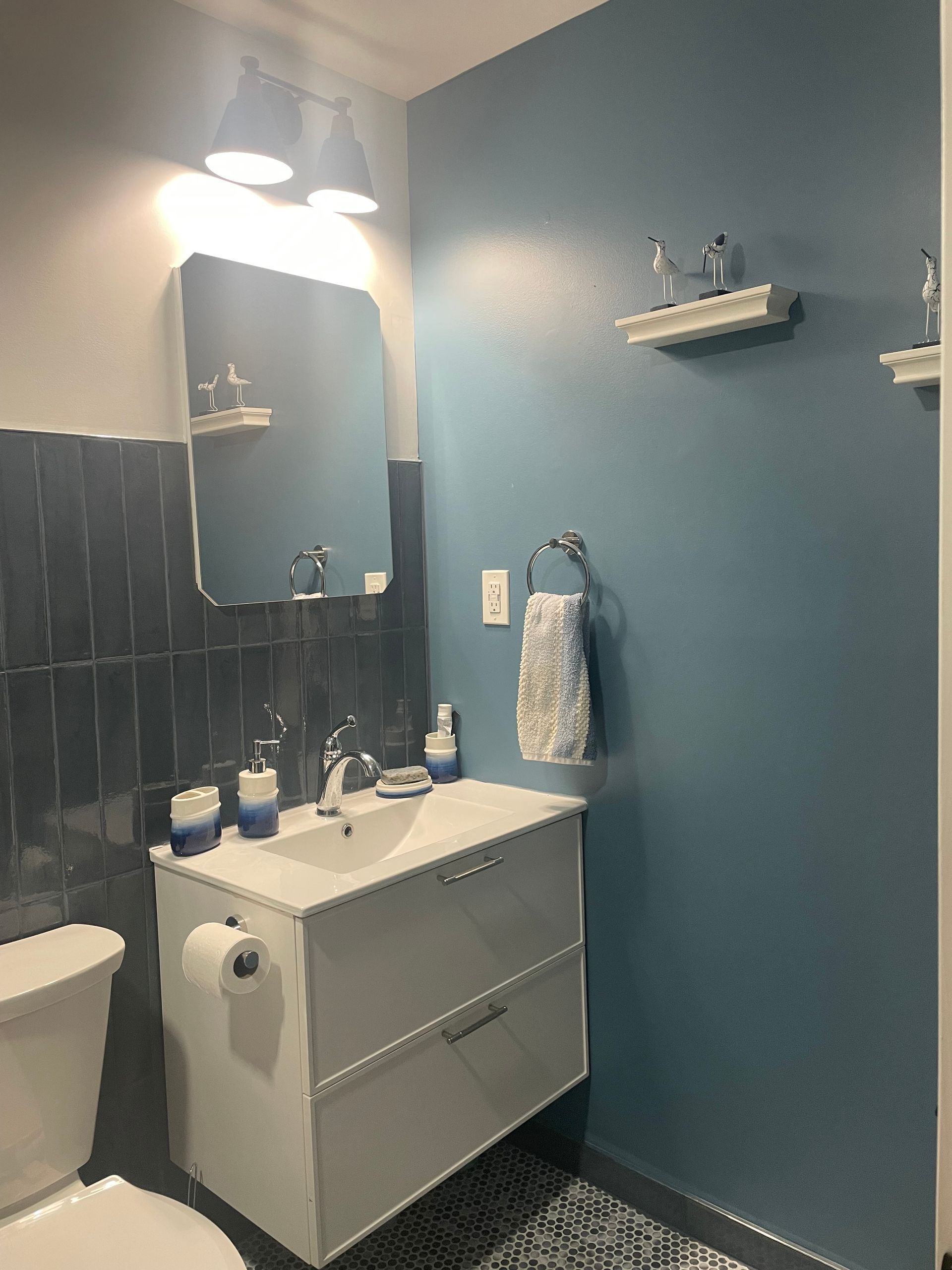 Blue bathroom vanity wall with mirror, wall shelves, light fixture, and gray tile wainscoting