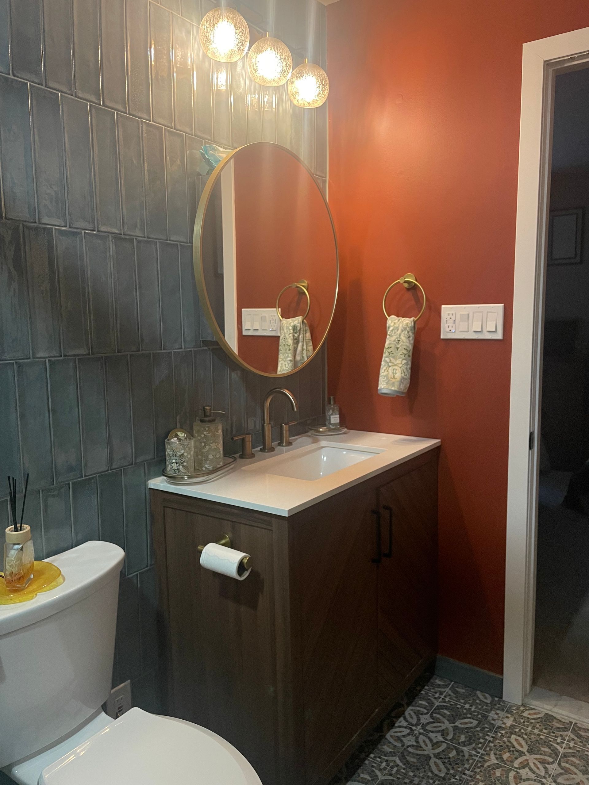 Bathroom vanity with oval mirror, globe lights, gray tile wall, and warm orange accent wall