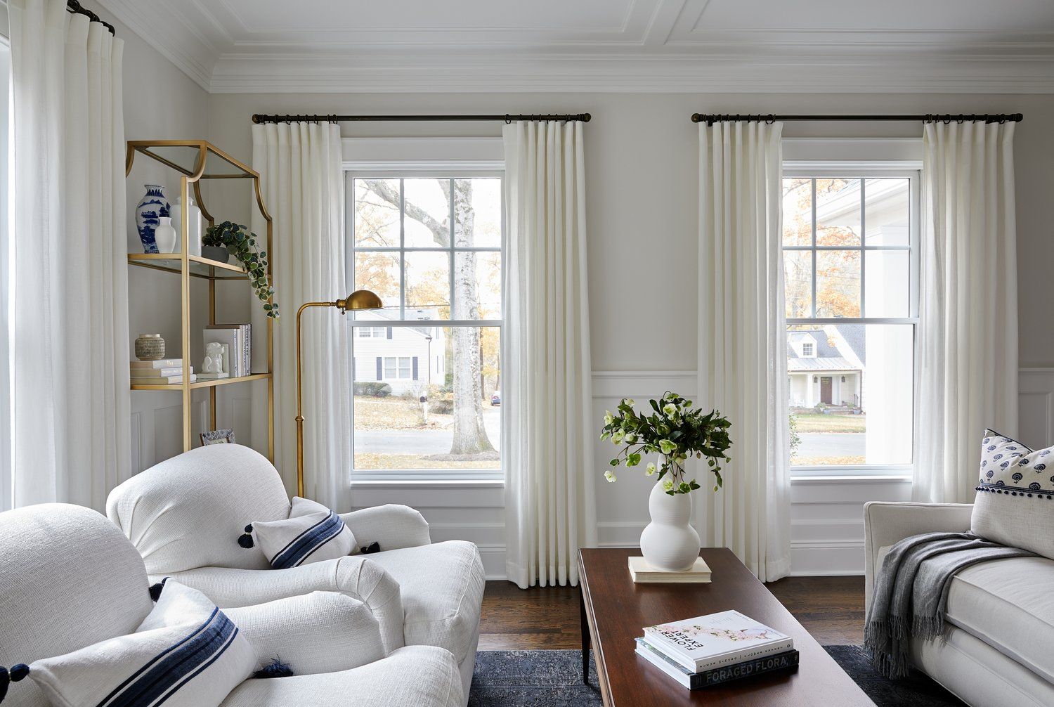 Bright living room with white sofa, tall windows, gold shelving, and soft neutral décor