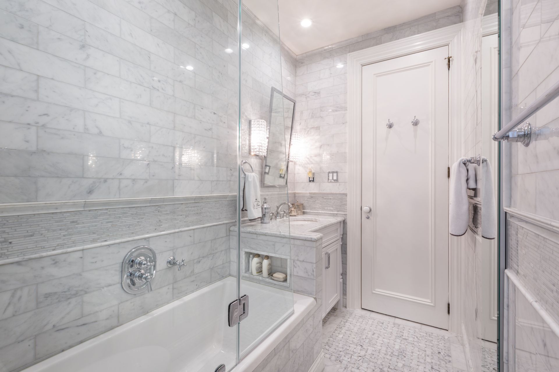 Narrow marble bathroom with glass shower, vanity, wall sconces, and white door