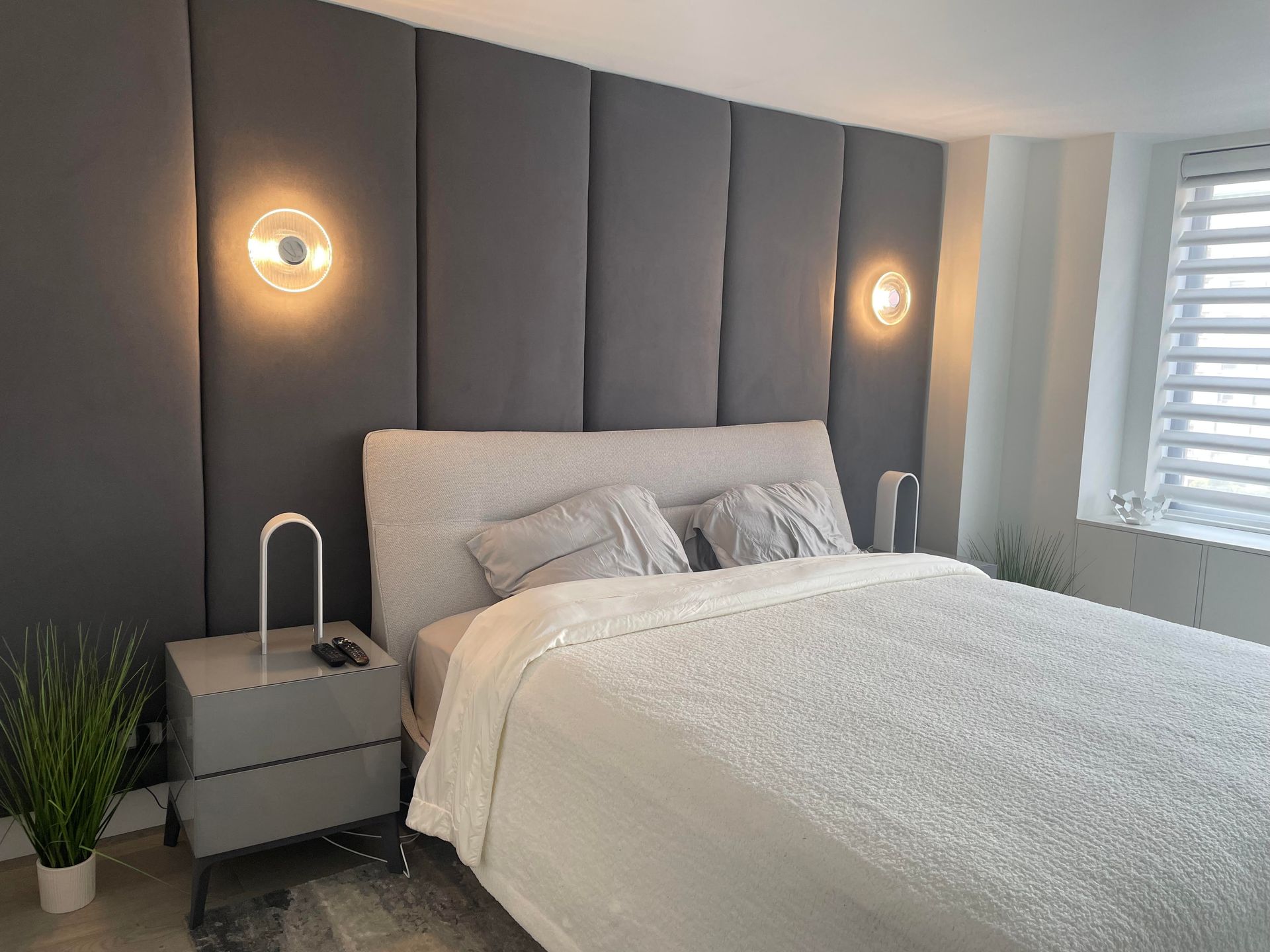 Modern bedroom with upholstered gray headboard, wall sconces, white bedding, and window shutters