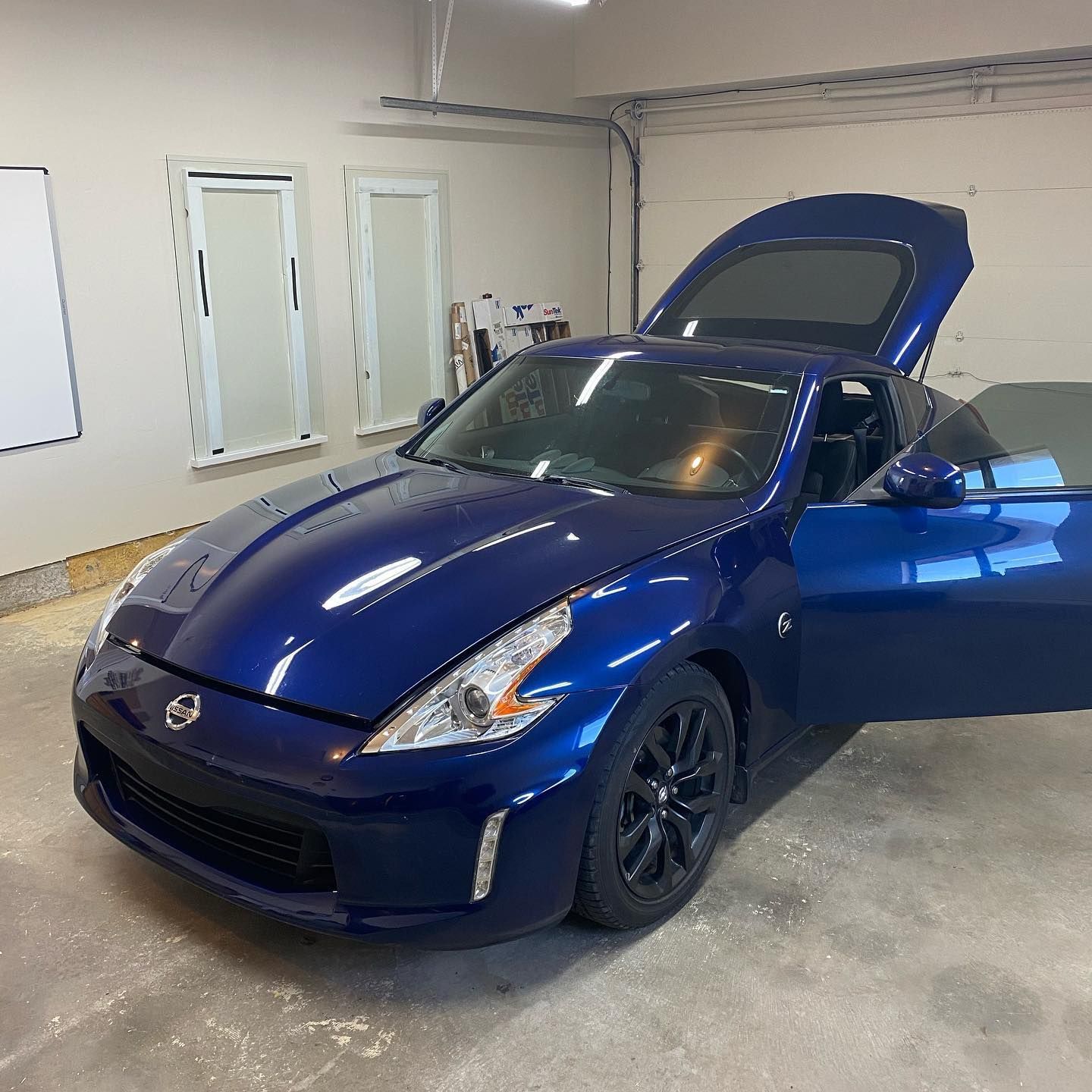 Blue Nissan 370Z sports car with the trunk and driver-side door open in a garage. Black wheels are visible.