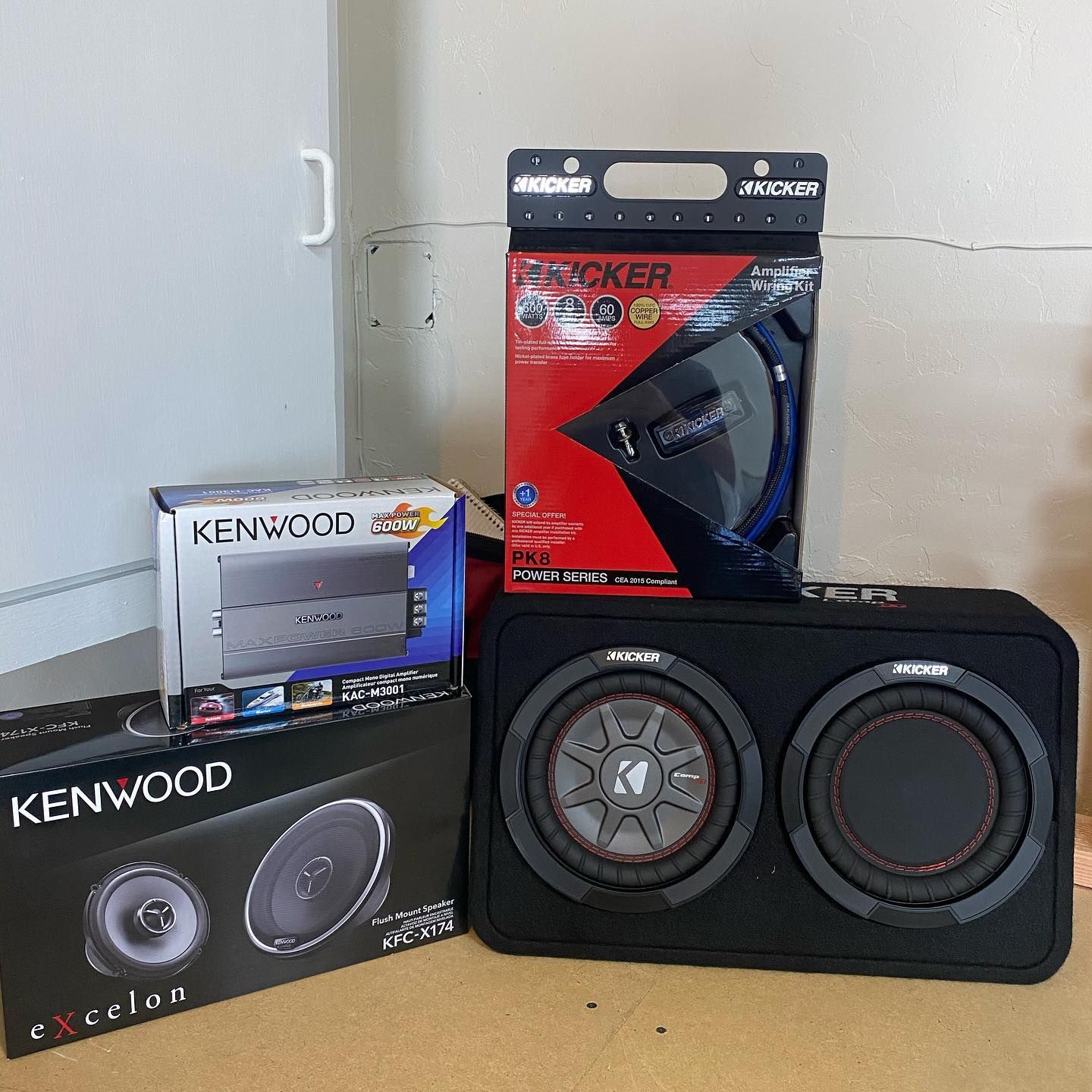 Car audio equipment including speakers, a subwoofer, and an amplifier are on a wood surface. A Kicker subwoofer box is front and center.