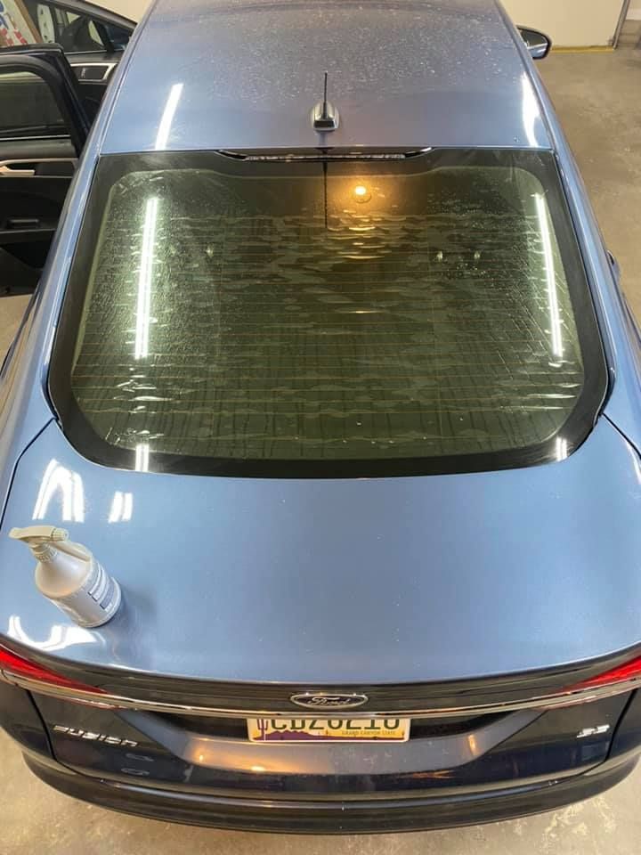 Blue sedan with rear window being cleaned. A spray bottle rests on the car's trunk.
