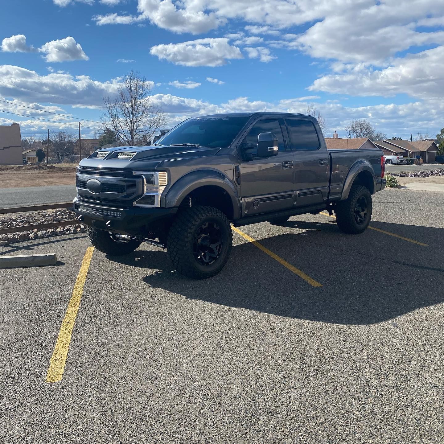 Dark gray lifted Ford pickup truck parked in a parking lot, with black rims and a sky background.