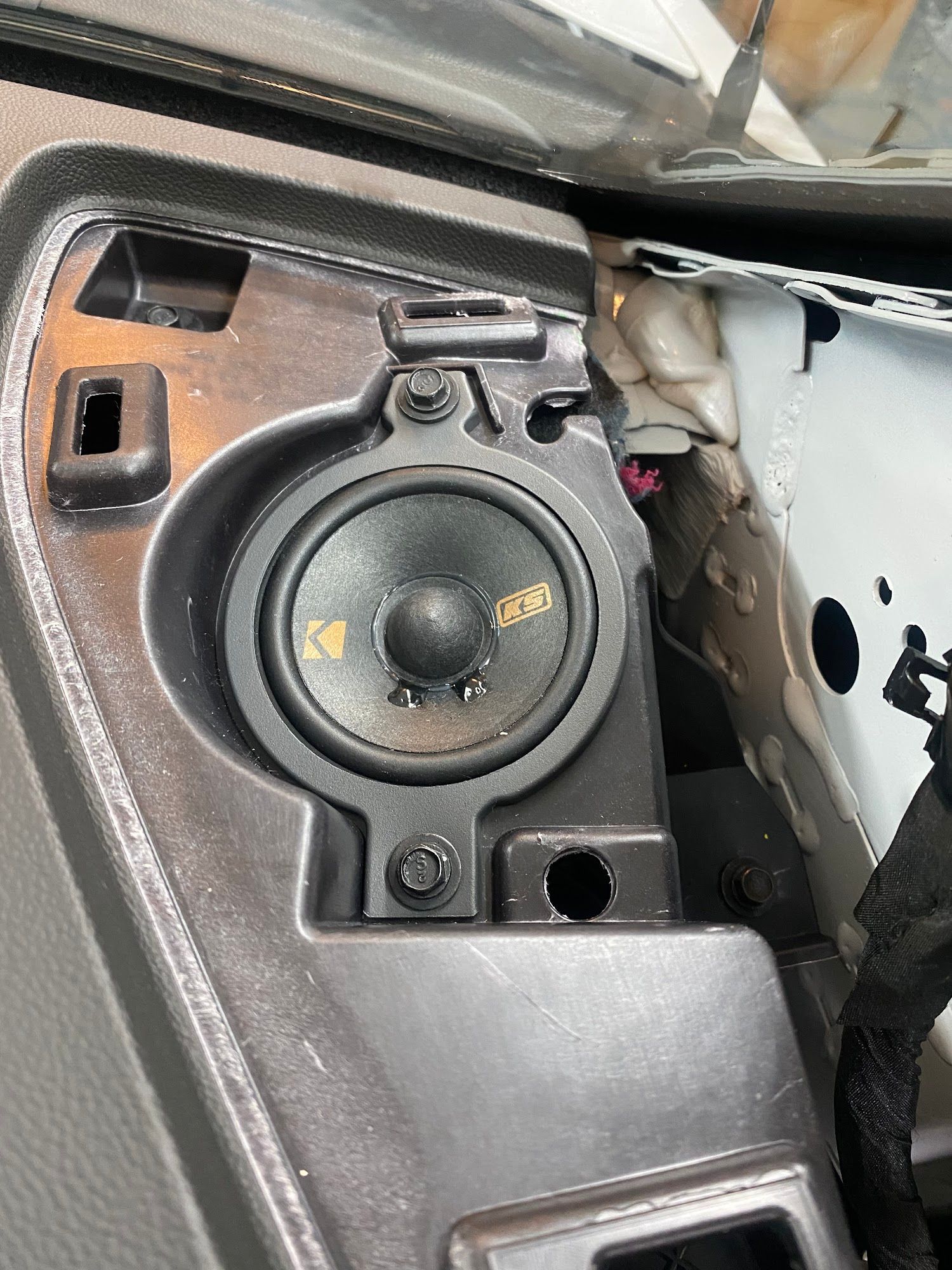 A car dashboard with a Kicker speaker installed. The speaker is black with a yellow logo.