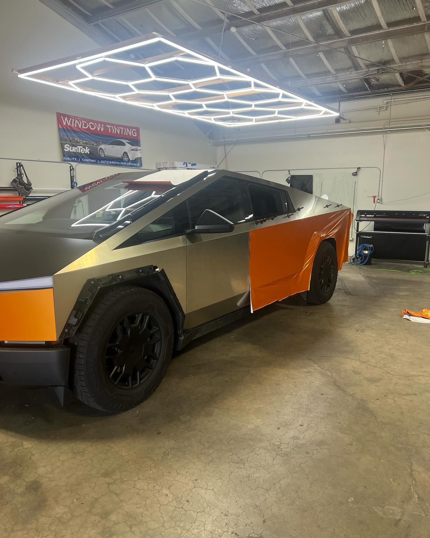 Tesla Cybertruck in a garage, partially wrapped in orange and gold vinyl. The car's front is black. Bright, geometric lights hang overhead.