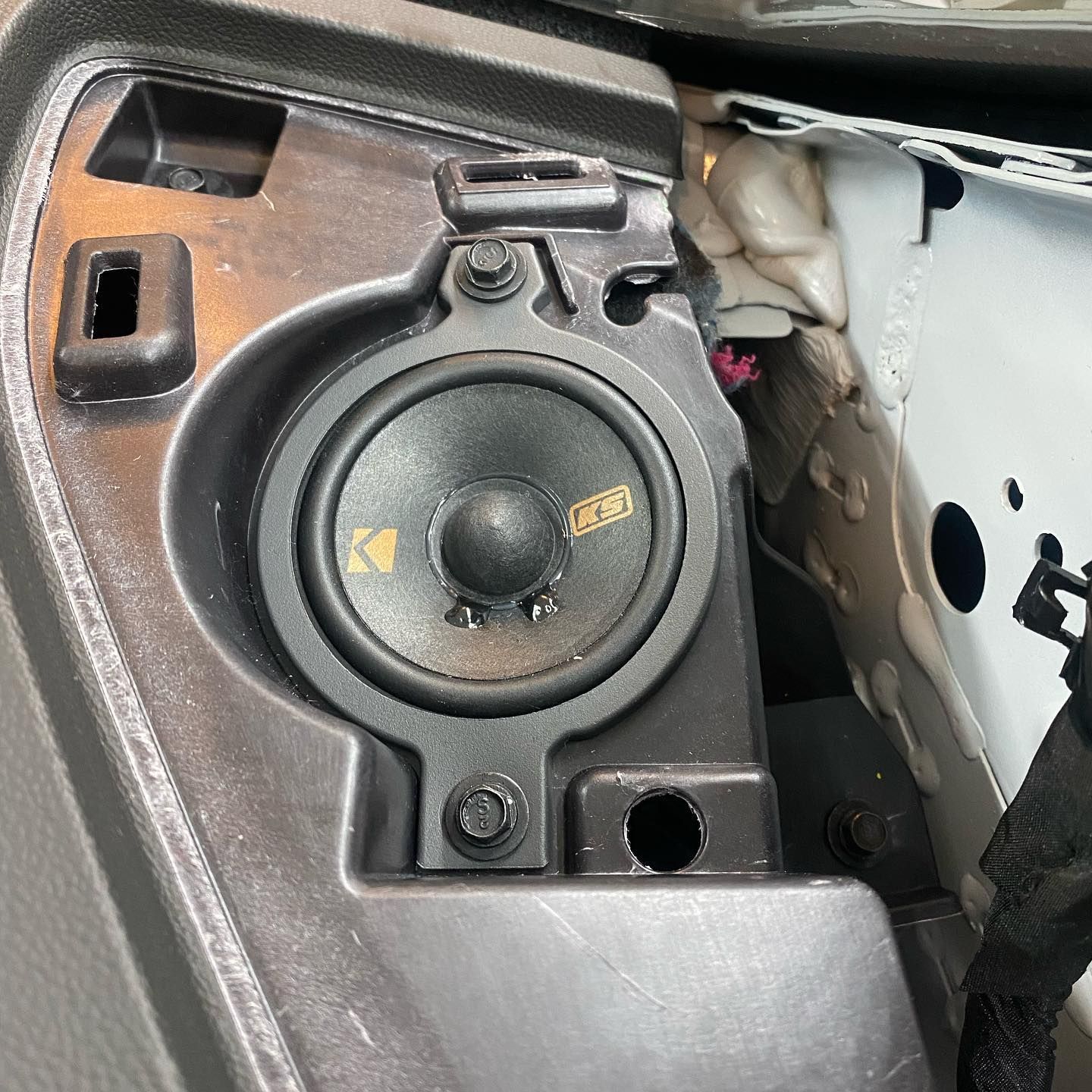 A close-up of a car's interior showing a black Kicker speaker mounted in a gray plastic dashboard panel.