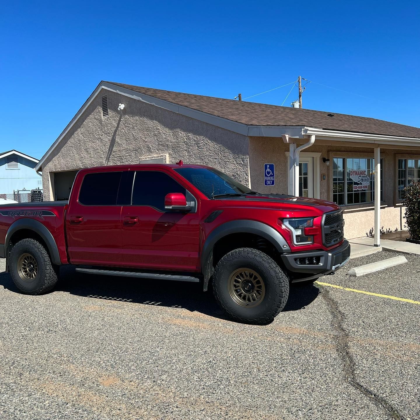 Red Ford Raptor truck parked in front of a beige building with a blue sky backdrop.