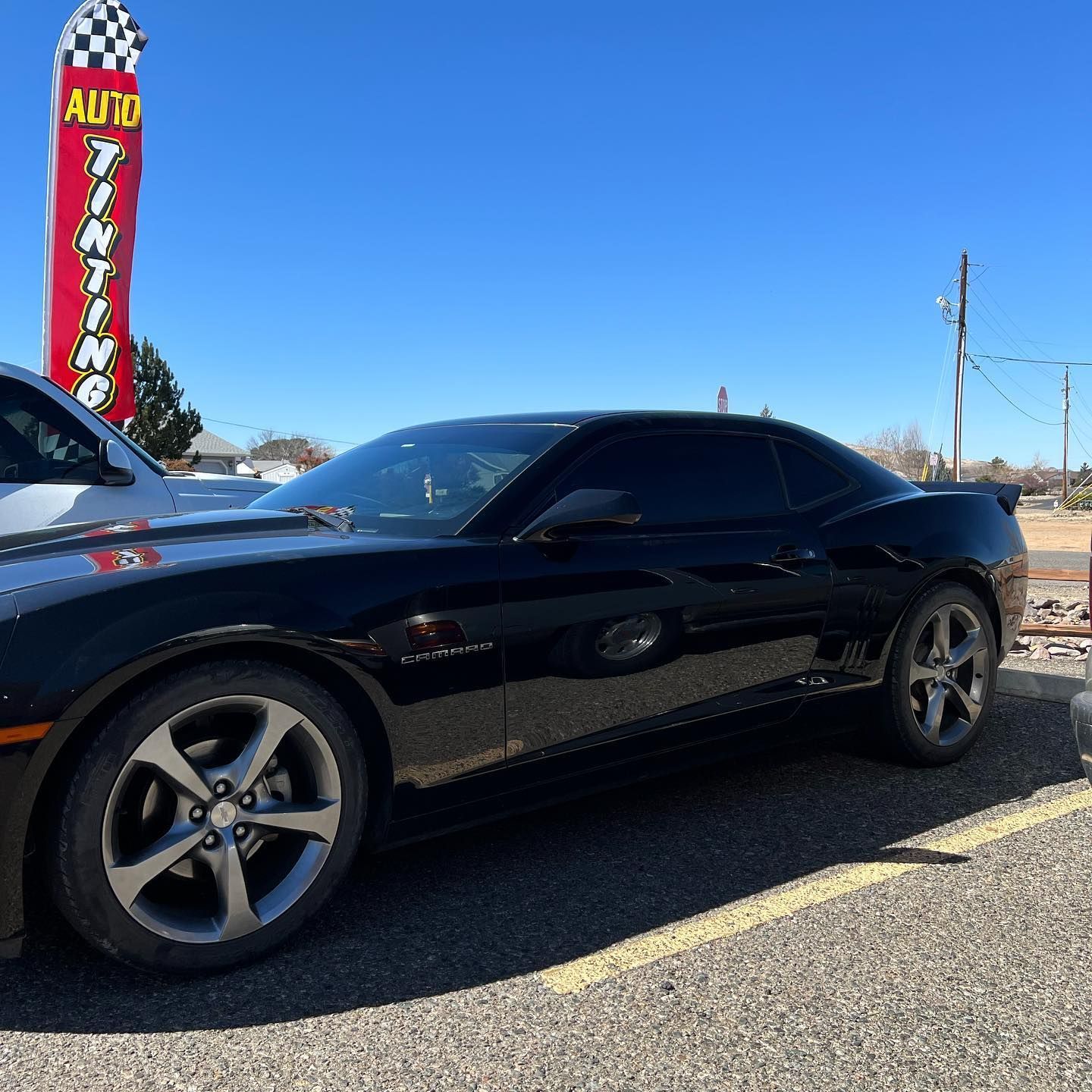 Black Chevrolet Camaro with tinted windows parked in front of an auto tinting shop on a sunny day.