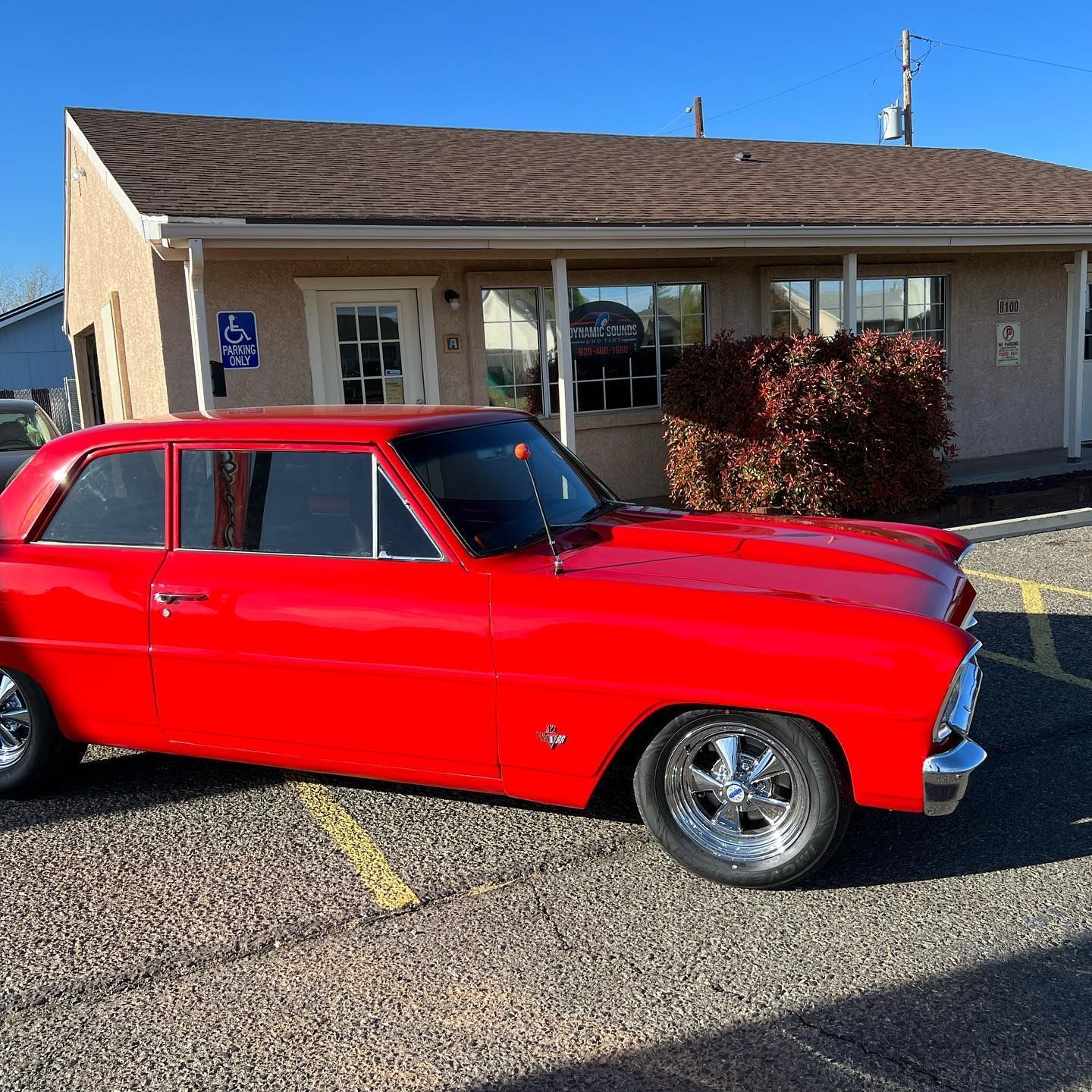 Red classic car parked in front of a light-colored building on a sunny day. The car has chrome wheels.