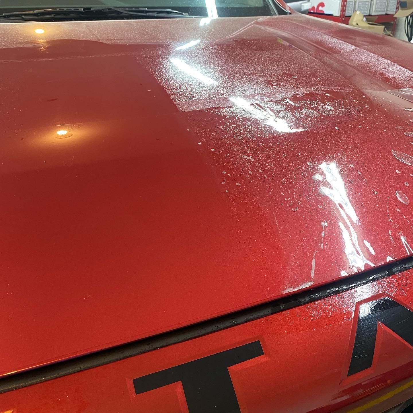 Red car hood with a wet, speckled surface, possibly from rain or a cleaning. Black lettering is visible on the hood.