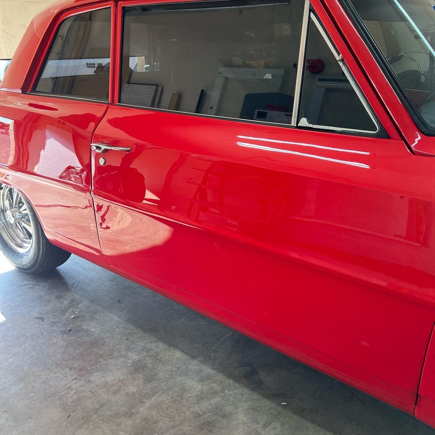 Red classic car parked on a gray concrete floor; side view showing doors, windows, and chrome wheels.
