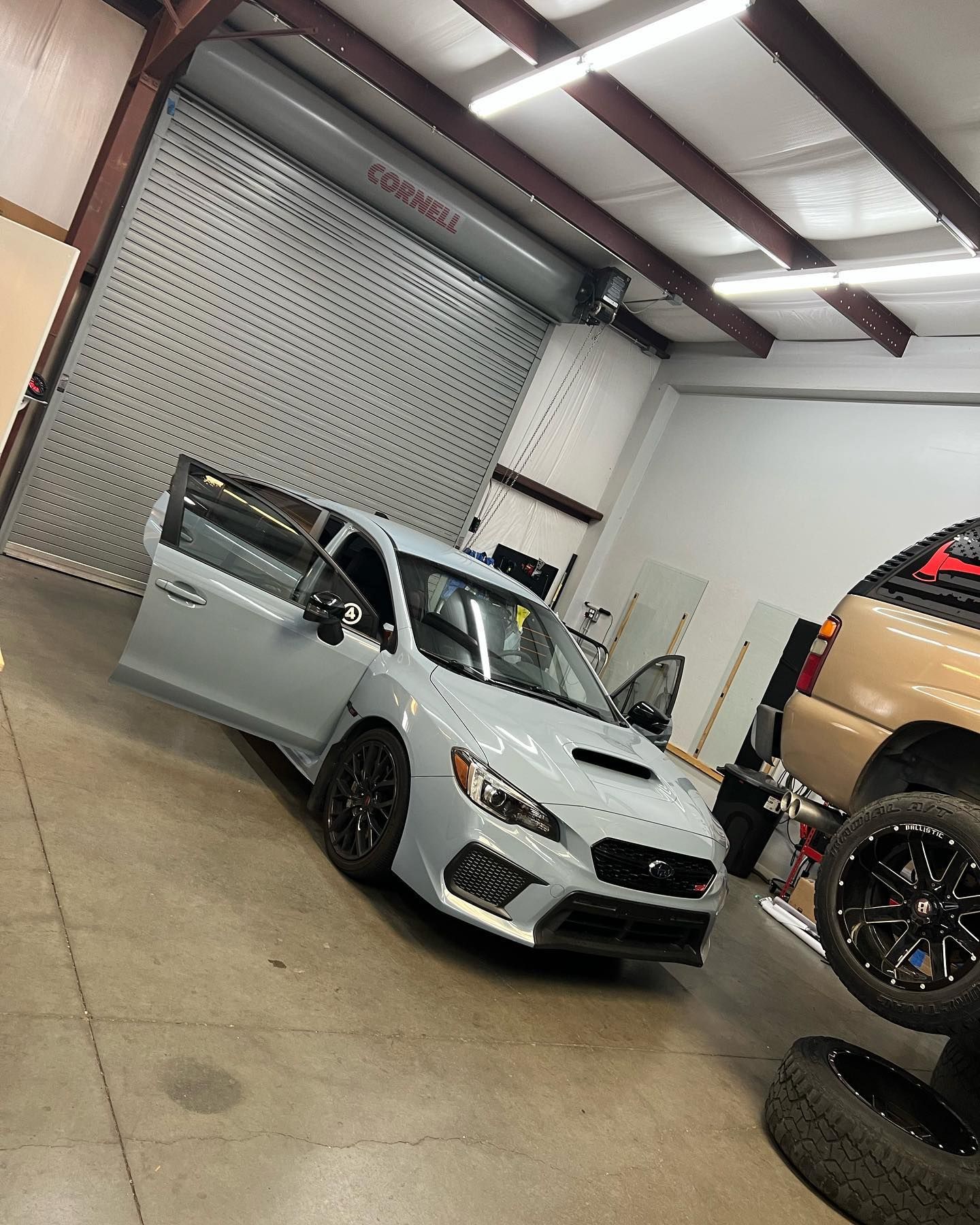 A light blue Subaru sports car with open doors inside a garage. A tan SUV is parked next to it.