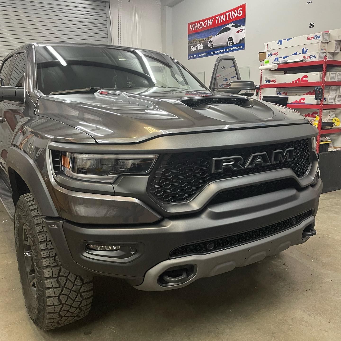 Dark gray Ram TRX pickup truck parked inside a shop, likely for window tinting. The truck has a black grille and off-road tires.
