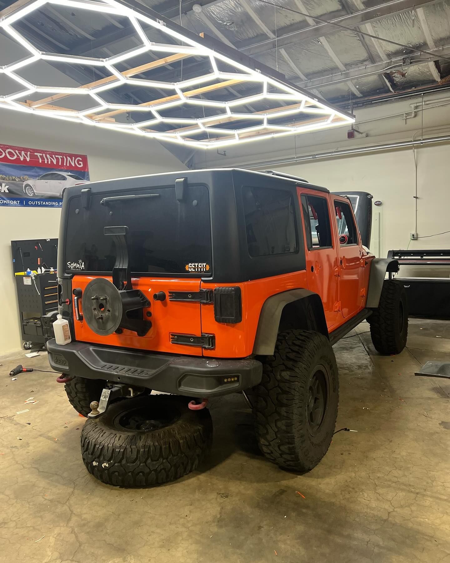 Orange Jeep Wrangler with black top and tires inside a garage, viewed from the rear. A spare tire rests on the ground.