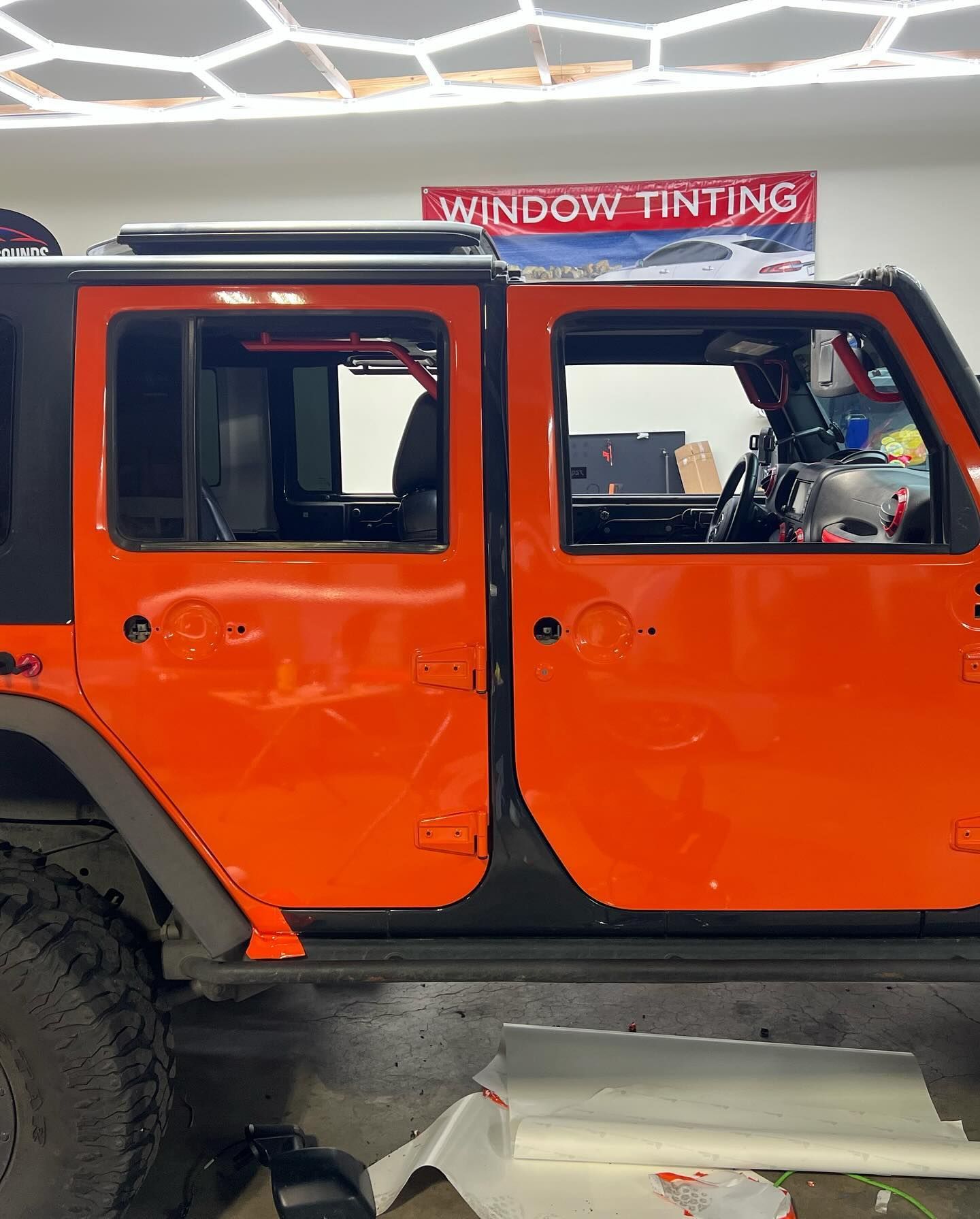 Bright orange Jeep Wrangler with open doors, ready for window tinting inside a shop. 