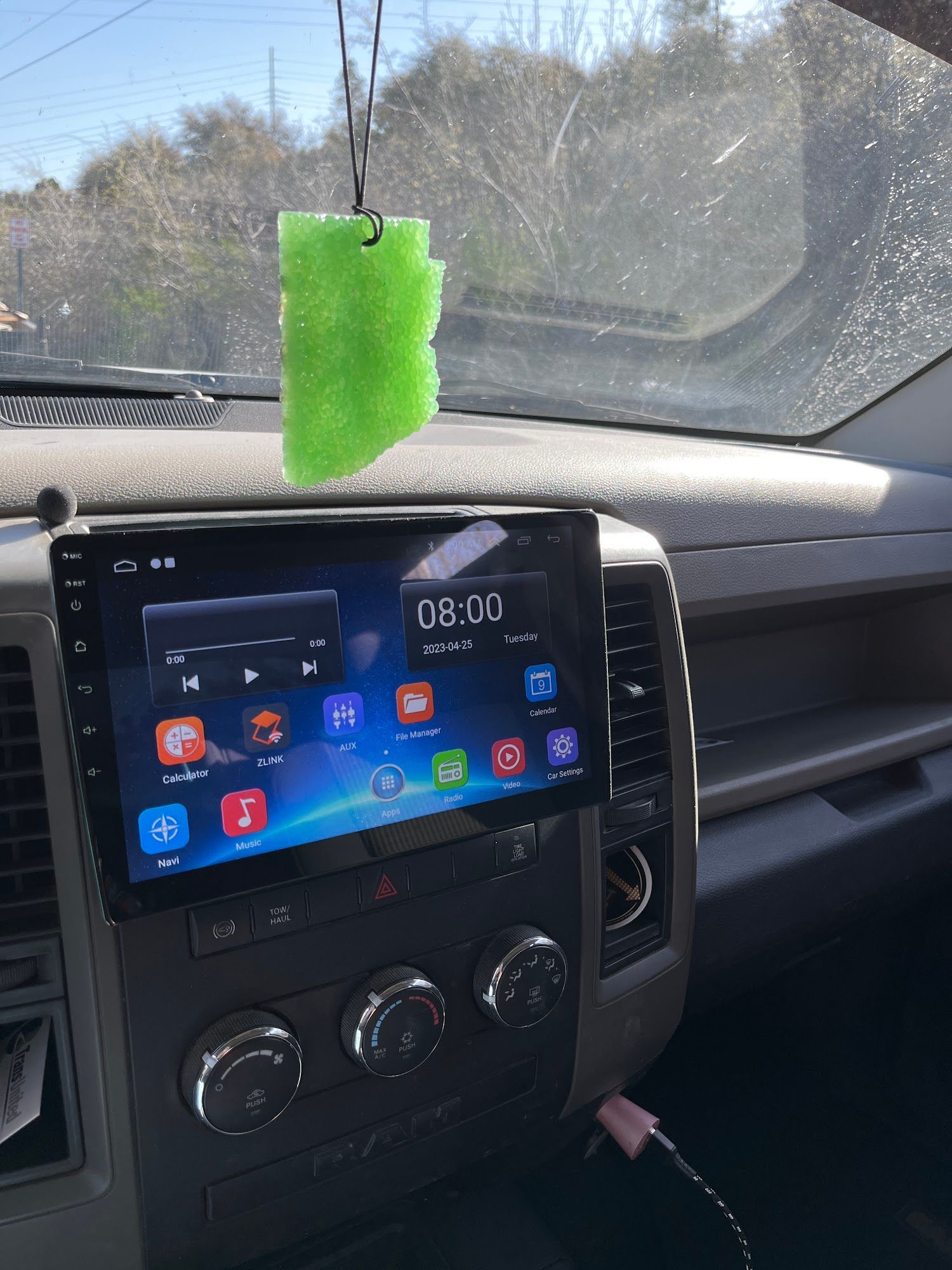 Inside a vehicle, a bright green air freshener hangs above a large touchscreen car stereo. The dashboard and controls are visible.