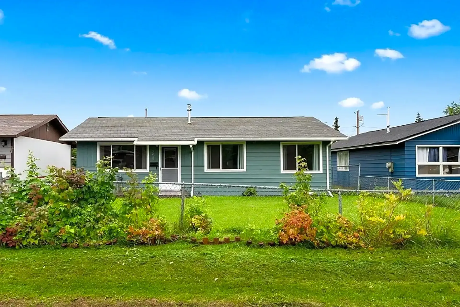 Blue house with a front lawn and small garden under a bright blue sky with some clouds.