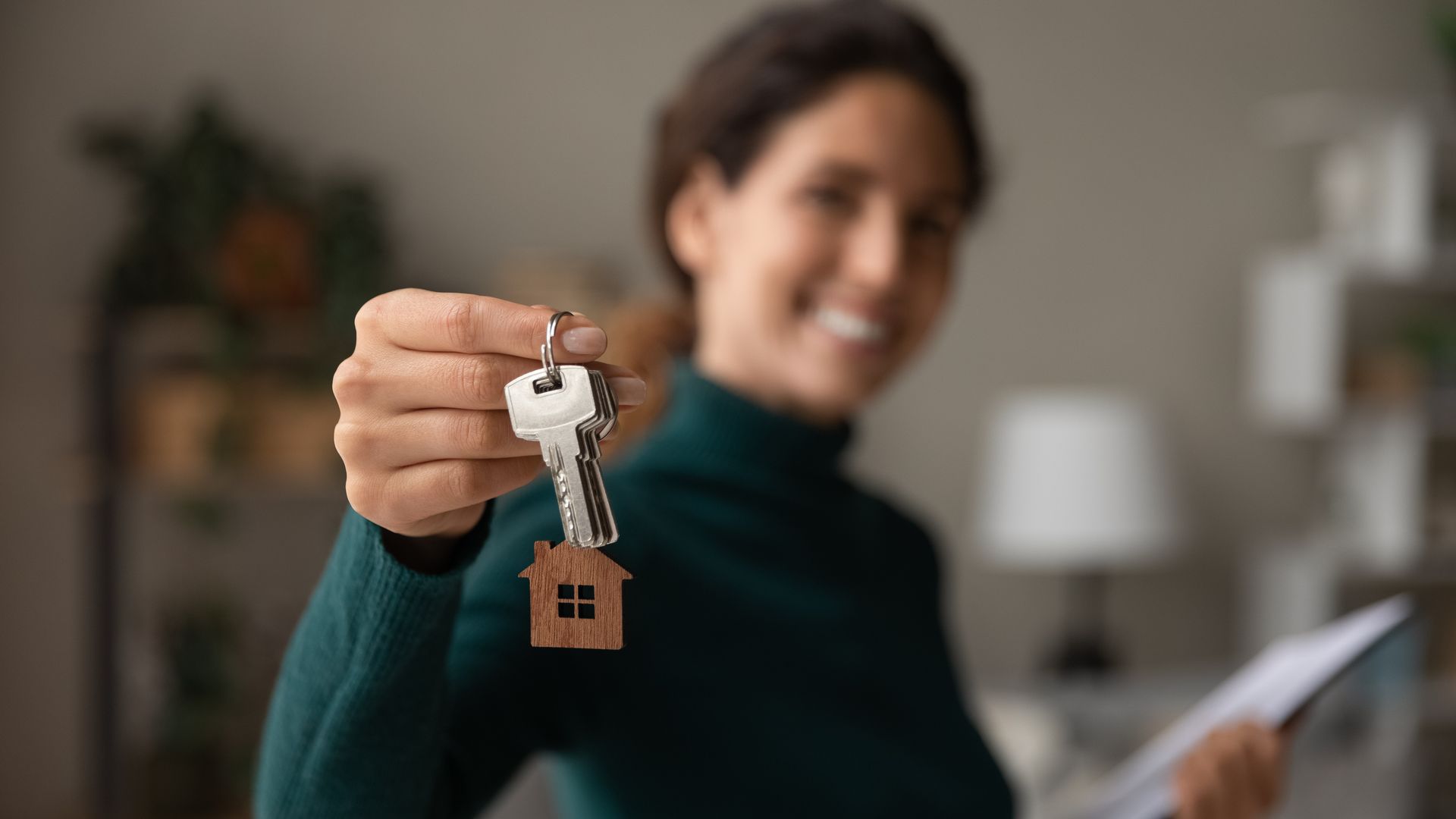 Person holding key with house-shaped keychain toward camera indoors.