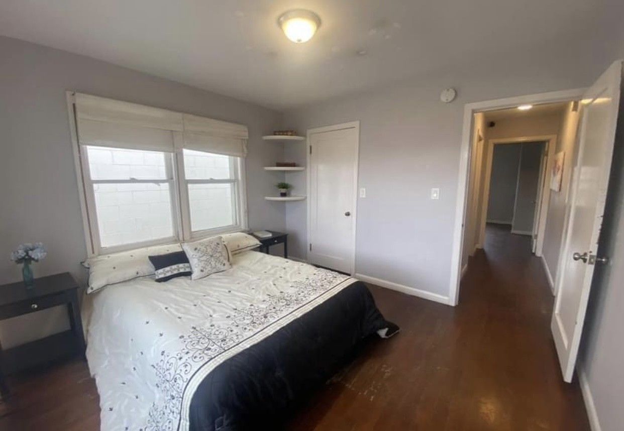 Bedroom with a bed, nightstand, closet, shelving, and hallway. The walls are grey, and floors are hardwood.