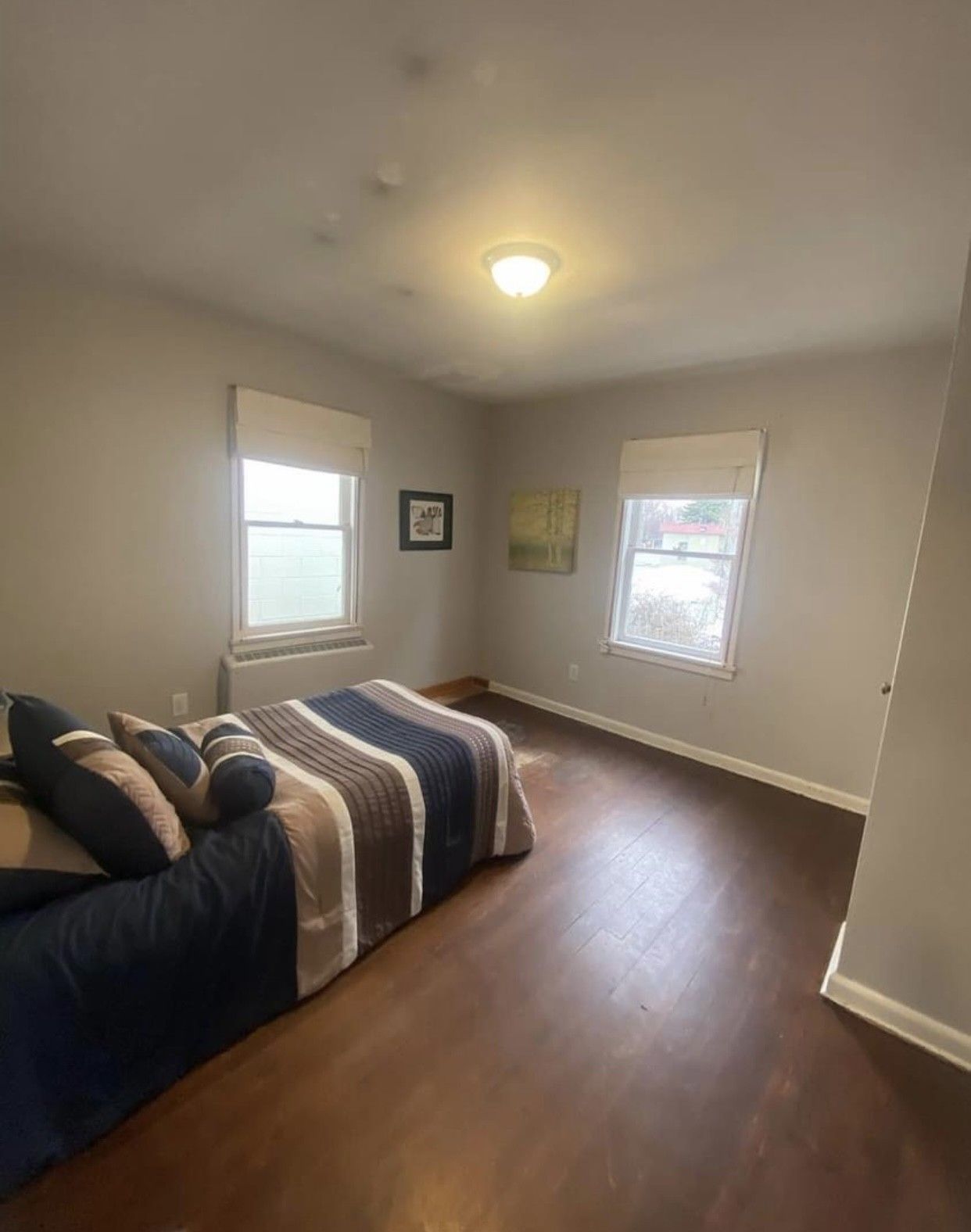 Bedroom with bed, two windows with shades, and dark wood flooring. Neutral walls and ceiling.