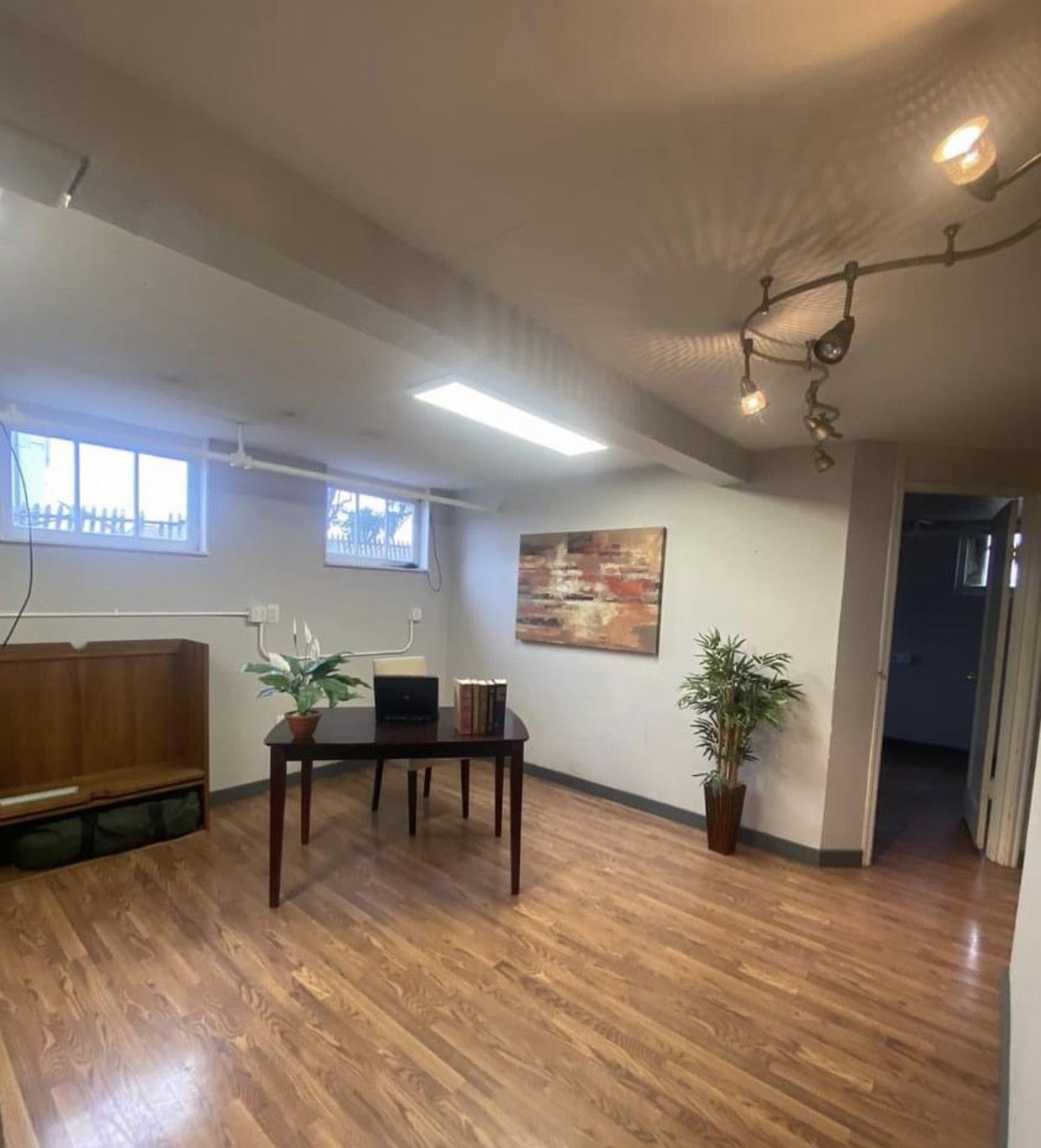 Basement office with a desk, art, and a plant. Wooden floor and white walls.
