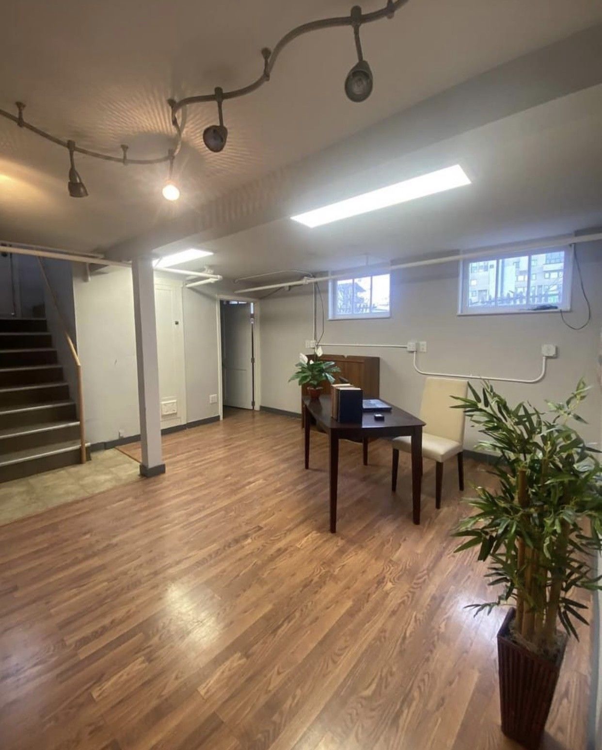 Basement office with wood floors, desk, chair, and plant. Stairs lead up on the left.