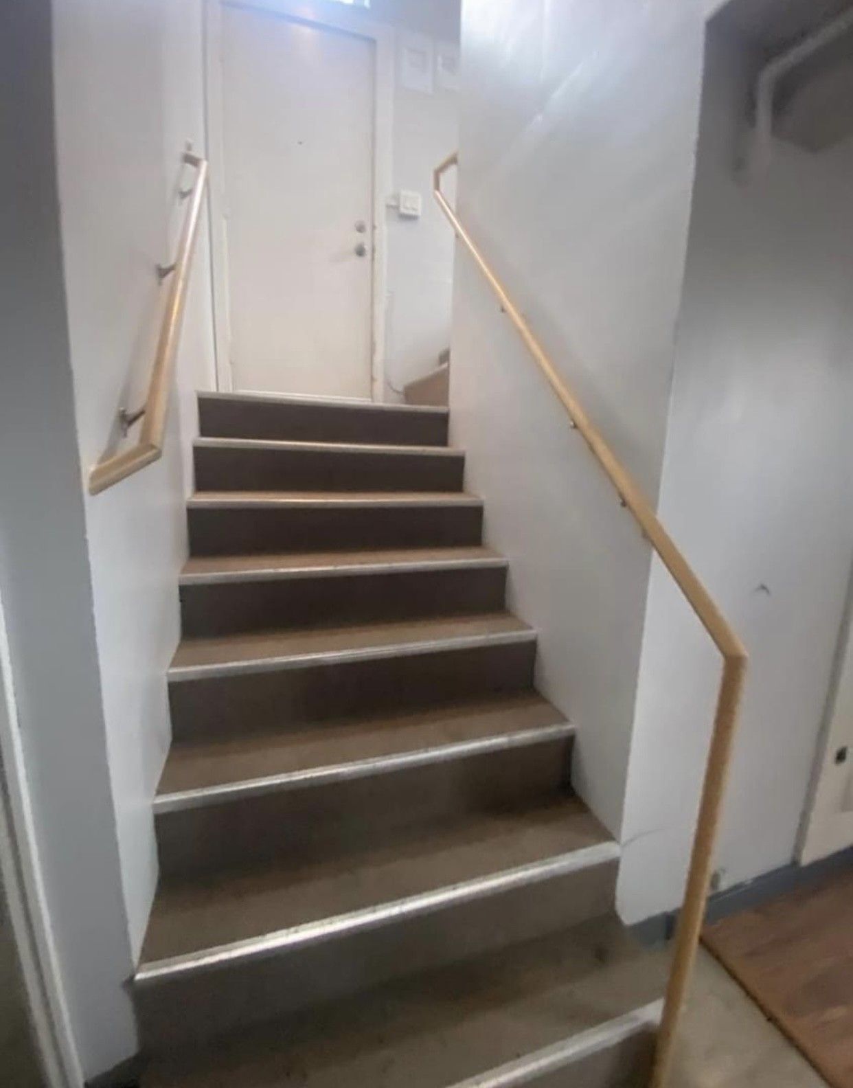 Staircase leading up to a closed white door. Brown carpet steps with light wood handrails. White walls.