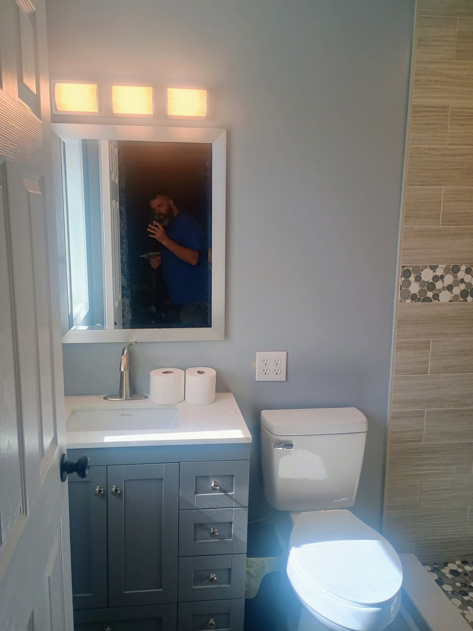 Small bathroom with blue vanity, mirror lights, toilet, and tiled shower wall; person reflected taking photo