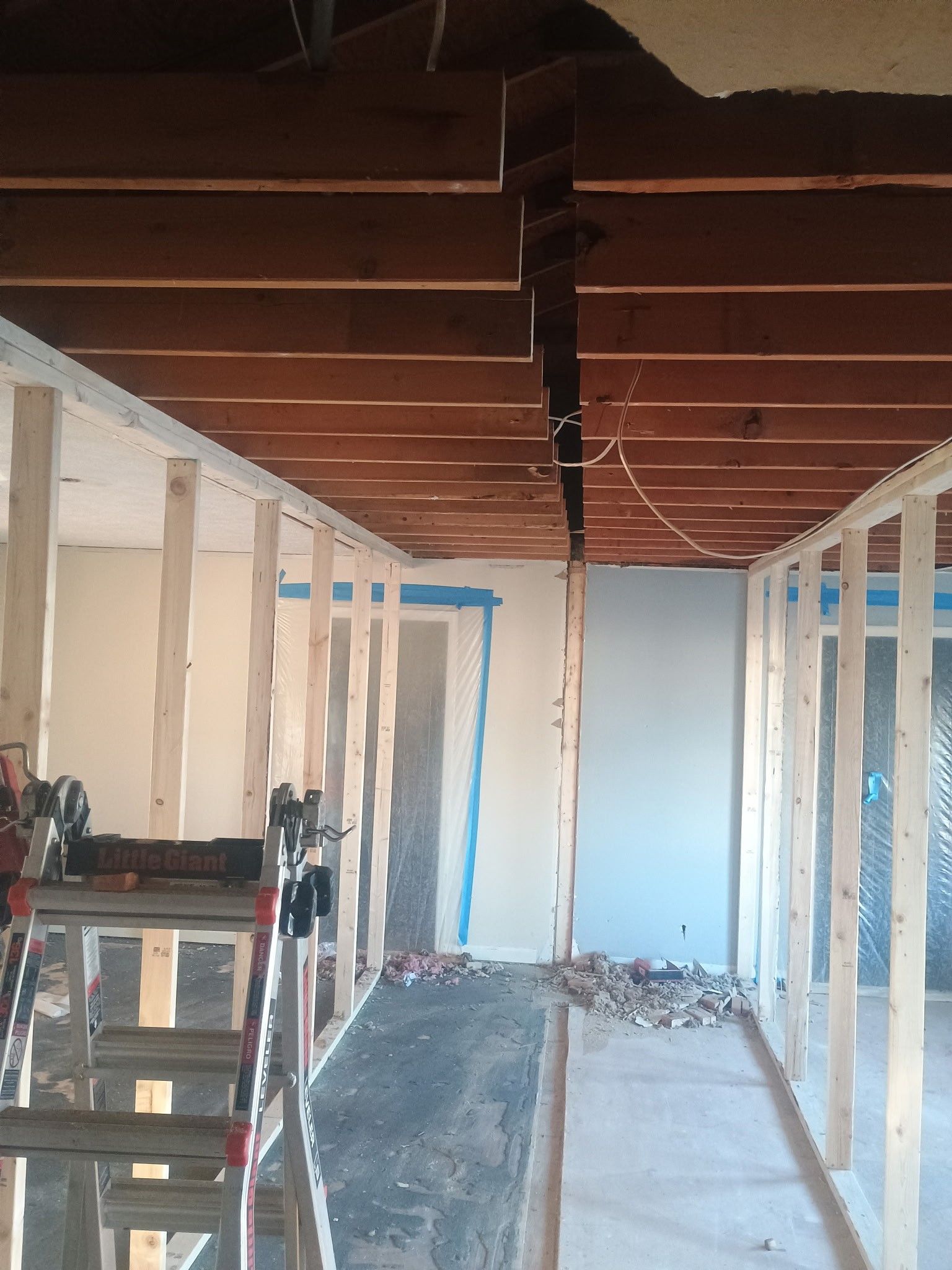 Interior framing under construction with exposed beams, white walls, and ladders on the left.