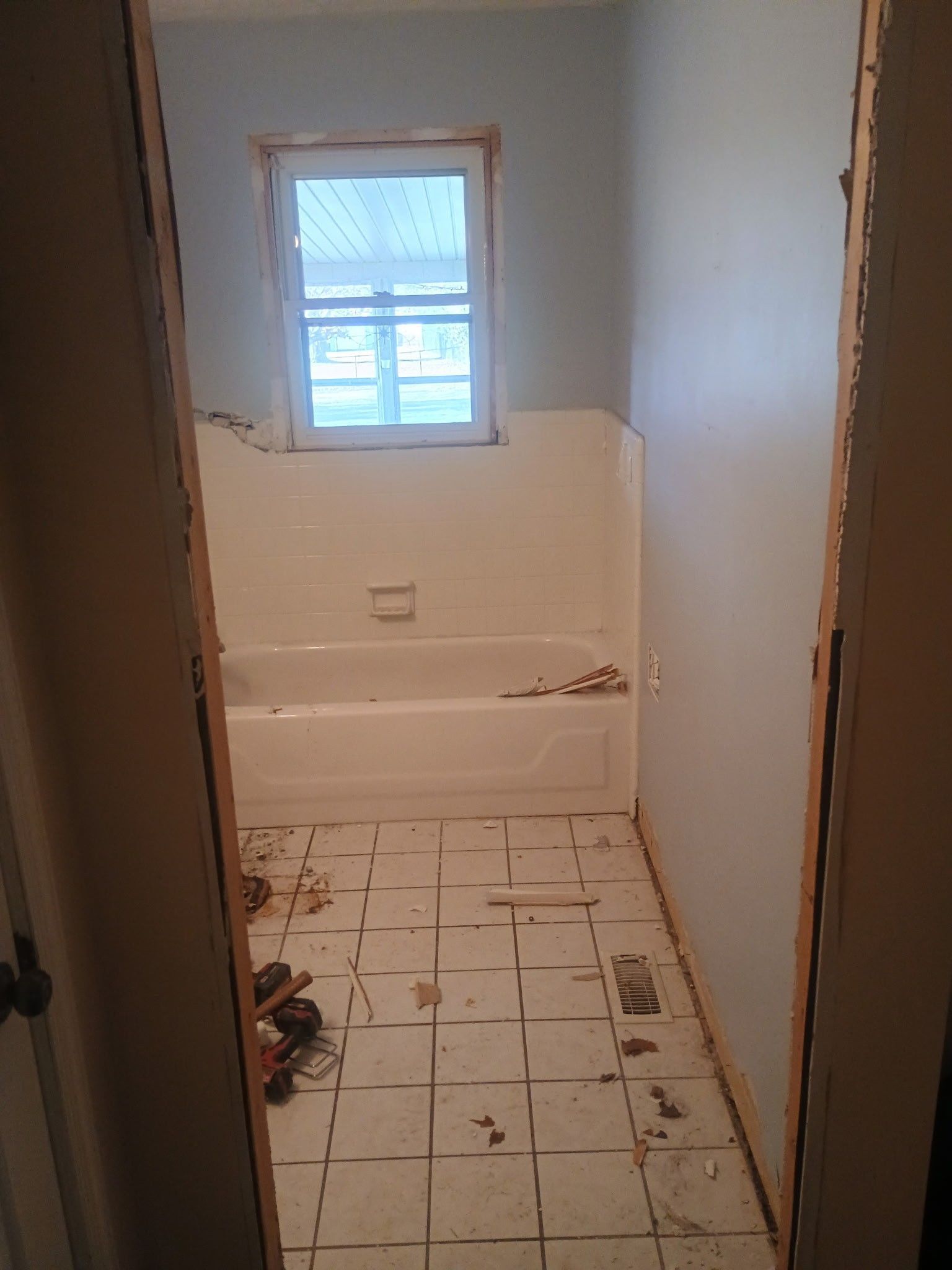 Small bathroom under renovation with white tub, exposed floor, and window with blue privacy covering.