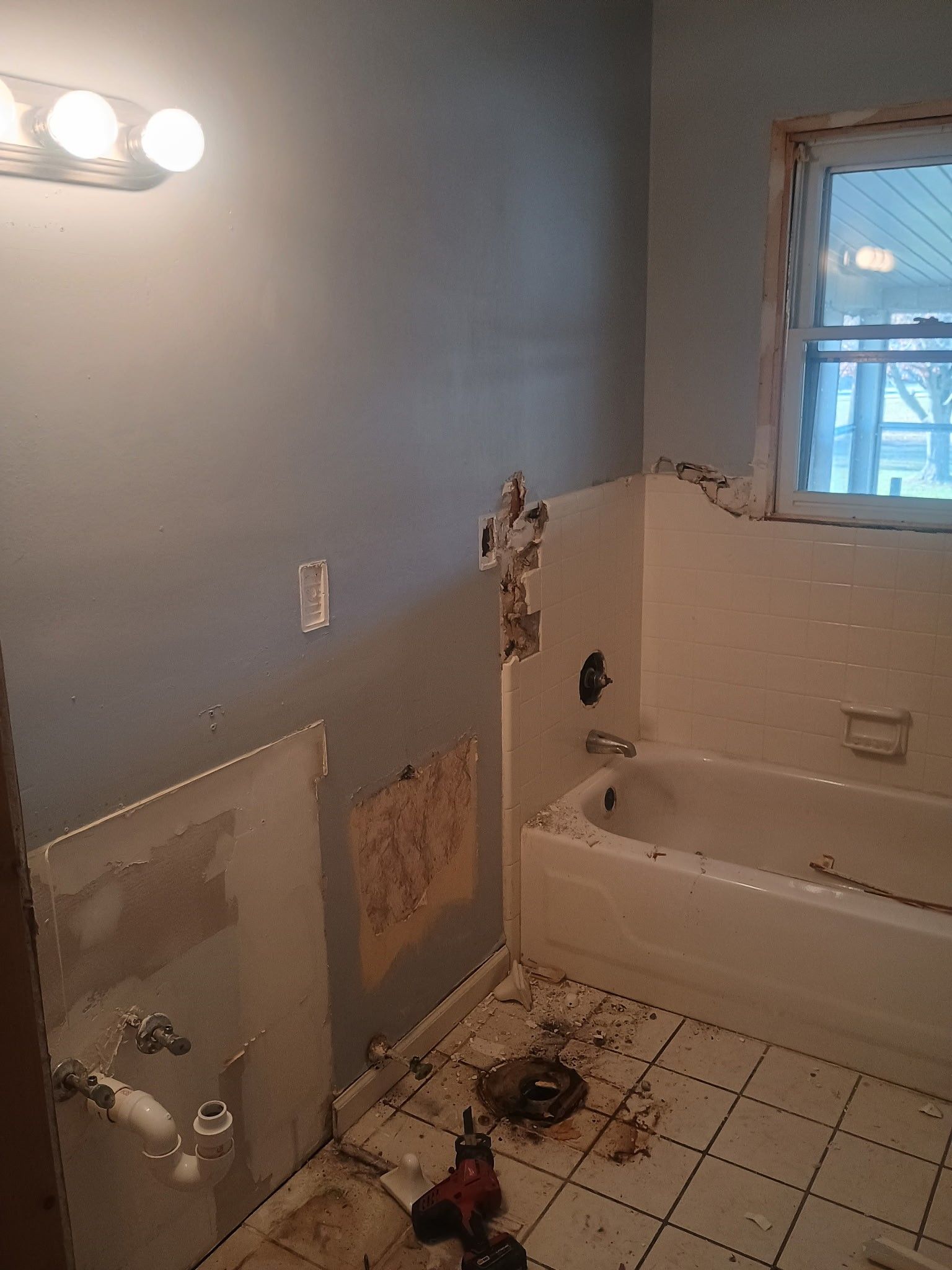 Damaged bathroom with torn drywall, debris on tile floor, and a bathtub by a window