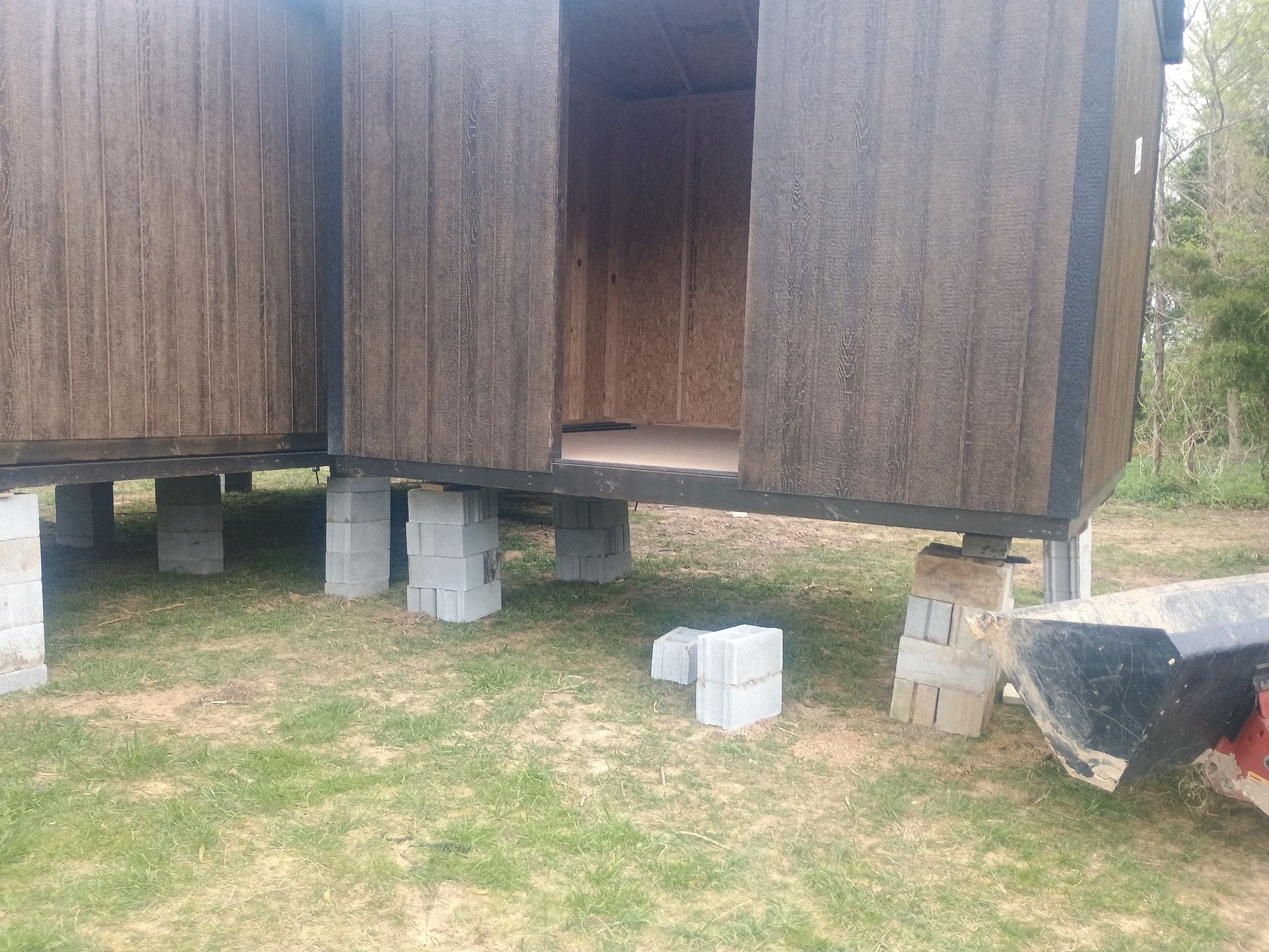 Elevated wooden shed on concrete blocks over patchy grass, with a dark object at right.