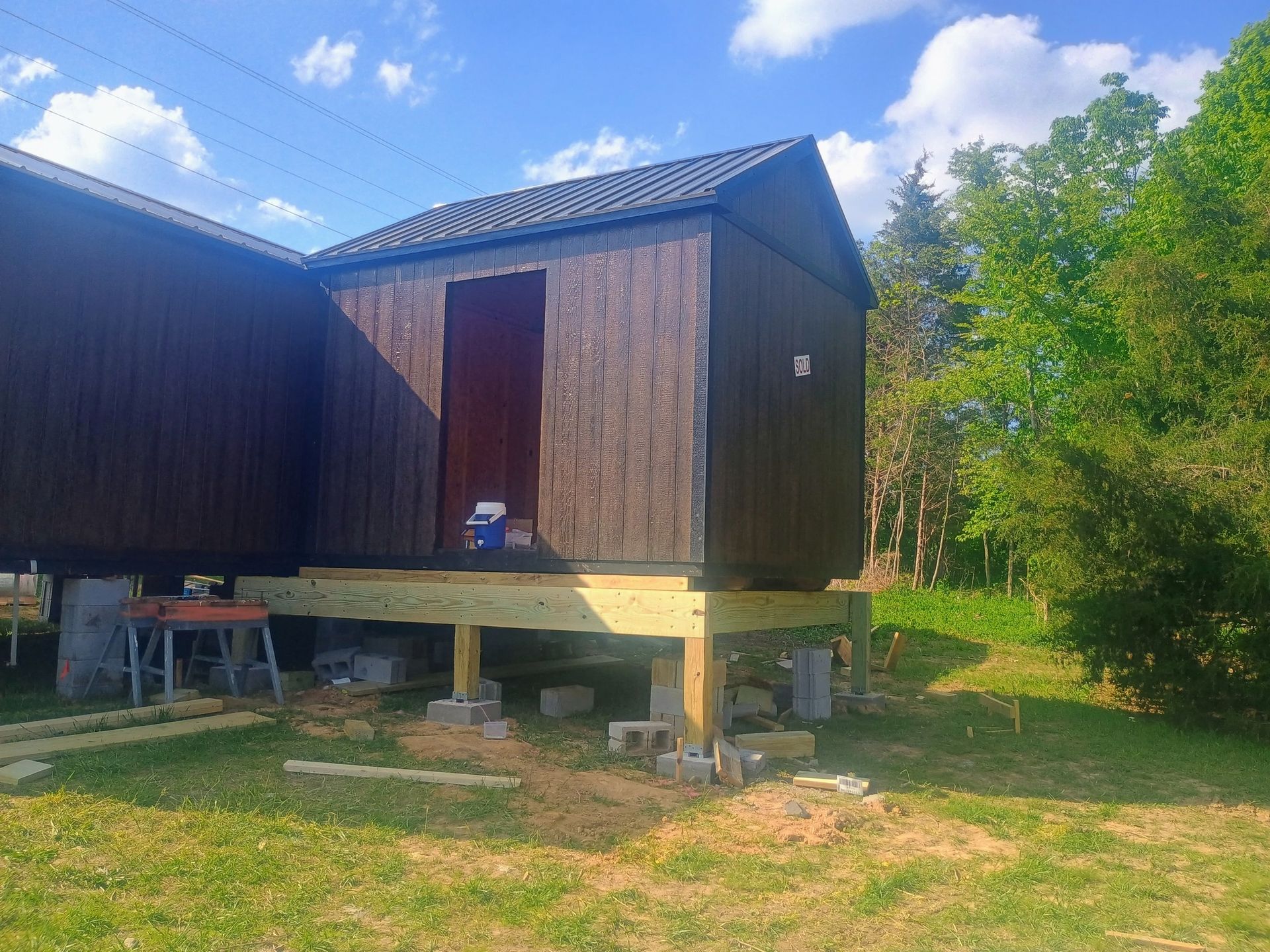 Wooden cabin on stilts with a red door, surrounded by grass and trees under a blue sky.