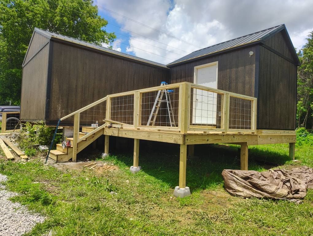 Two dark wood cabin-like buildings with a raised wooden deck and stairs in a grassy yard