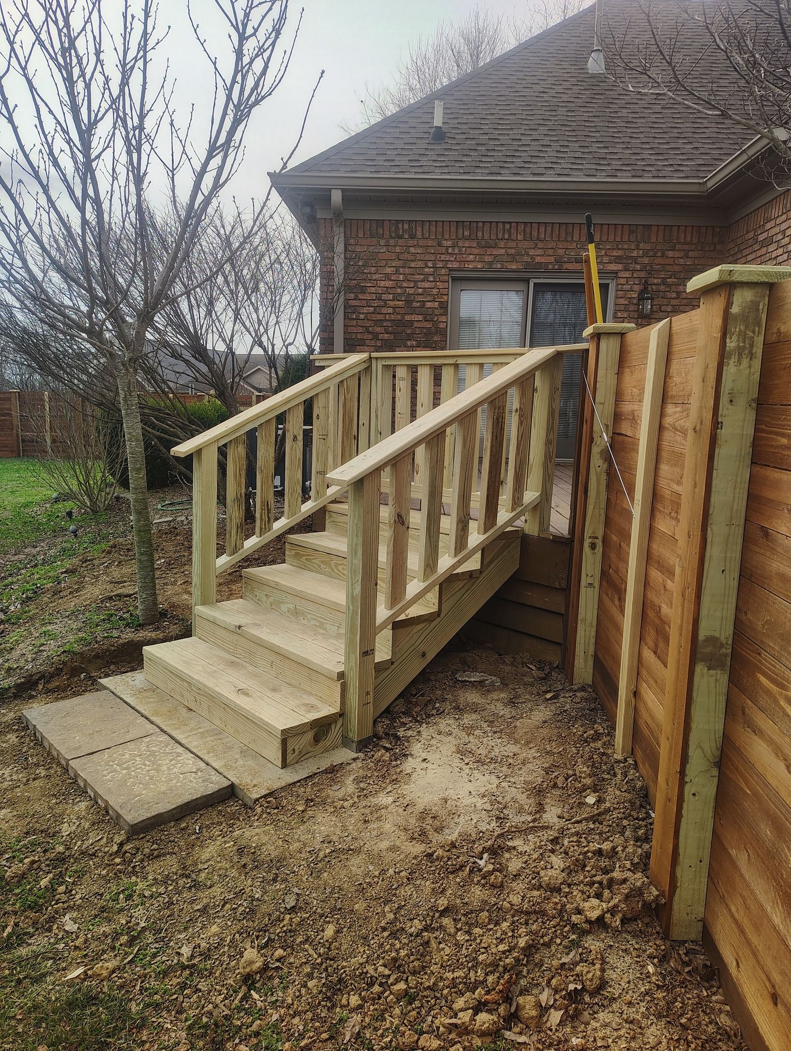 Wooden accessibility ramp with railings leading to a brick house entry, beside a fenced yard.