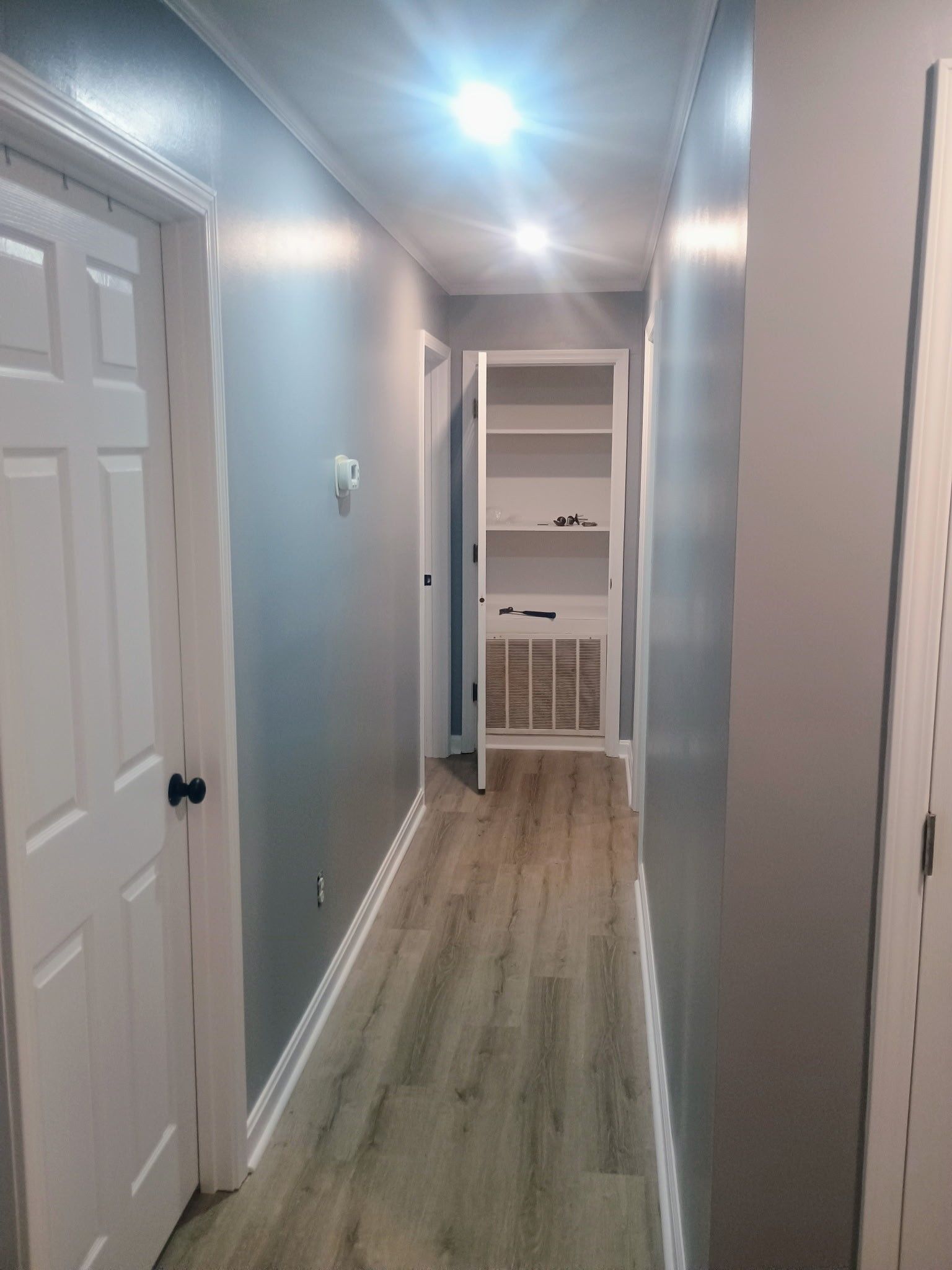 Narrow hallway with gray walls, white doors, and wood-look flooring leading to a small open room.