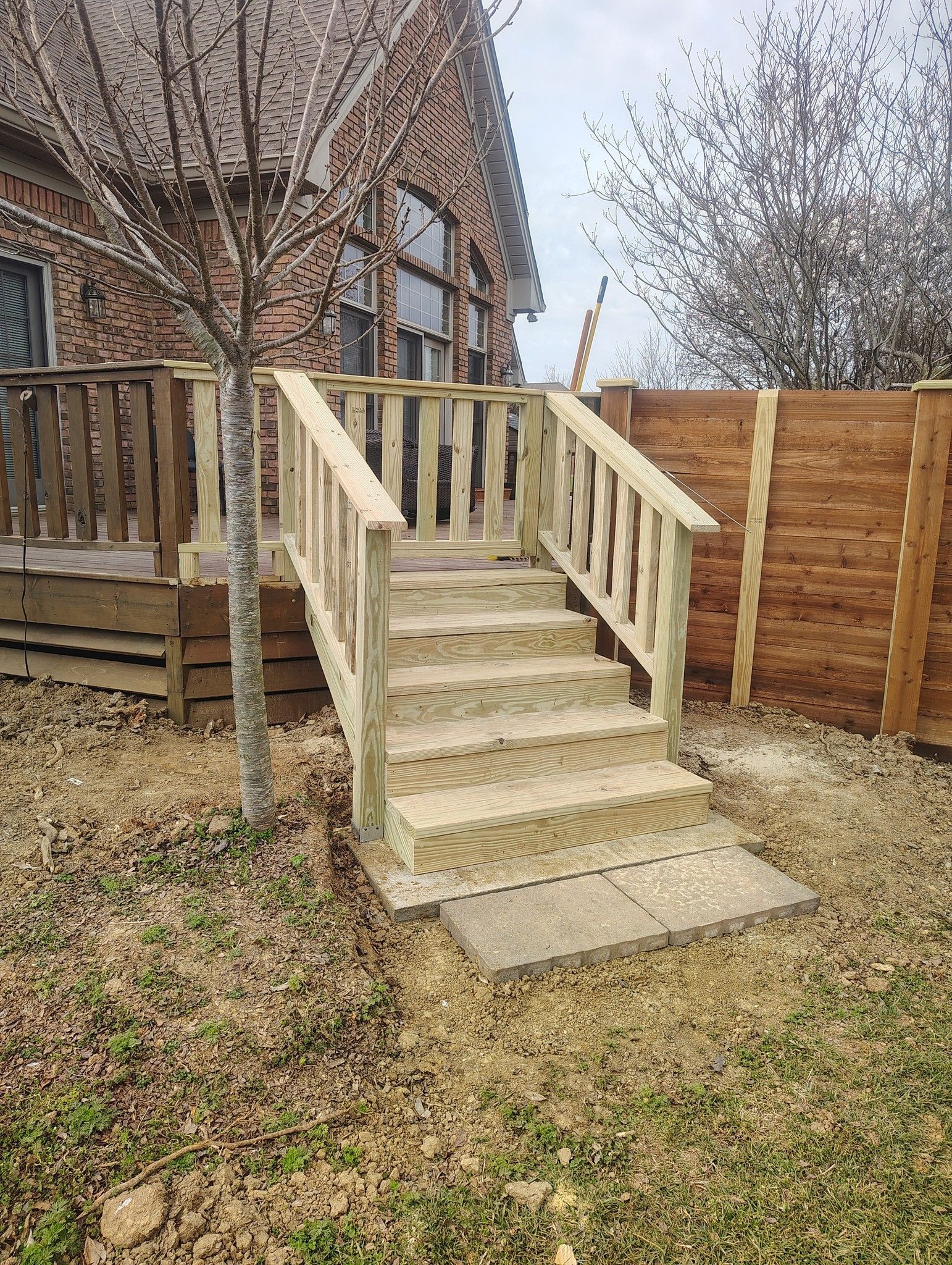 Wooden porch steps with railings leading to a house in a yard, bordered by a wooden fence.