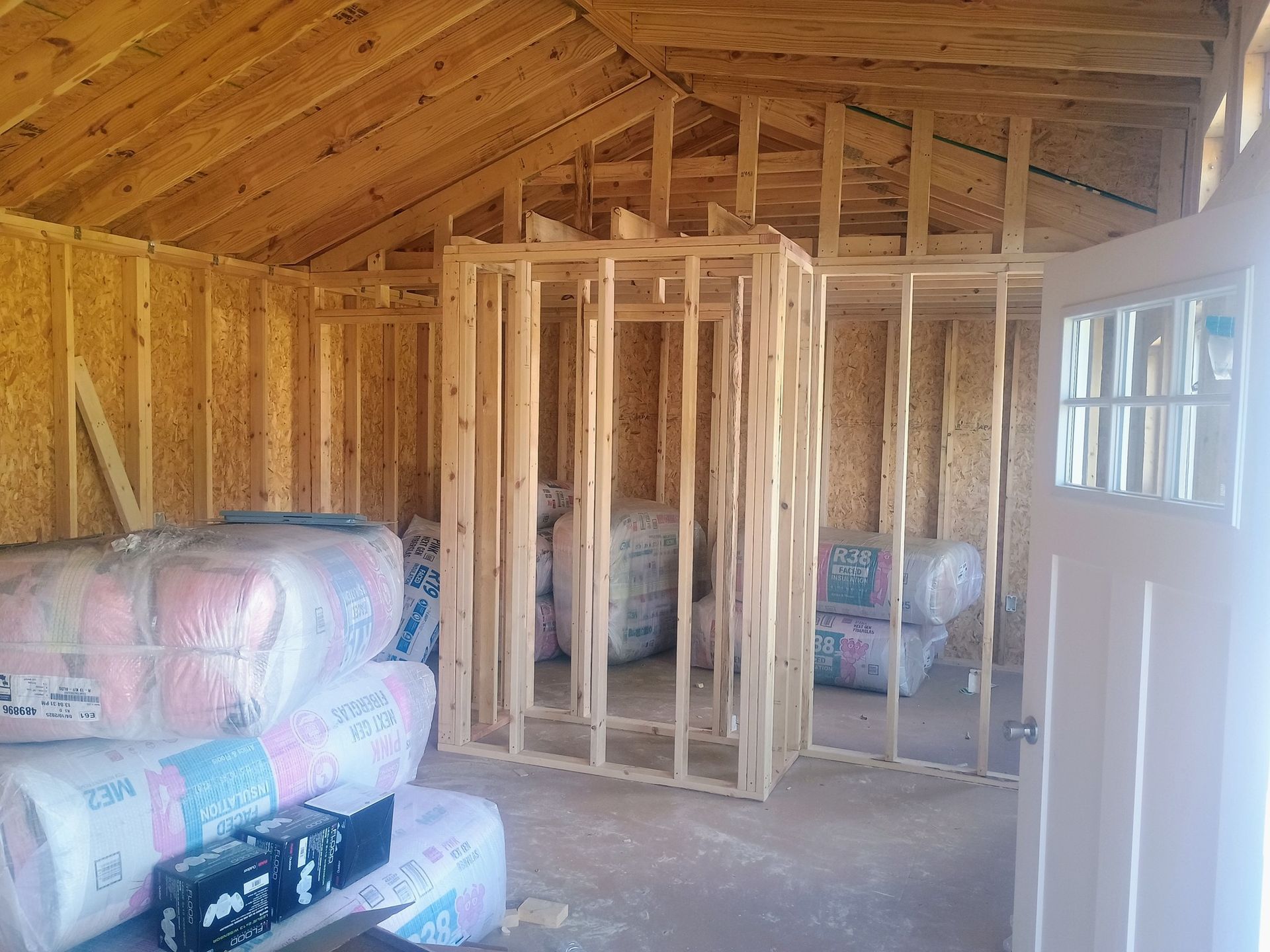 Unfinished attic room with exposed wood framing, insulation rolls, and a white door on the right.
