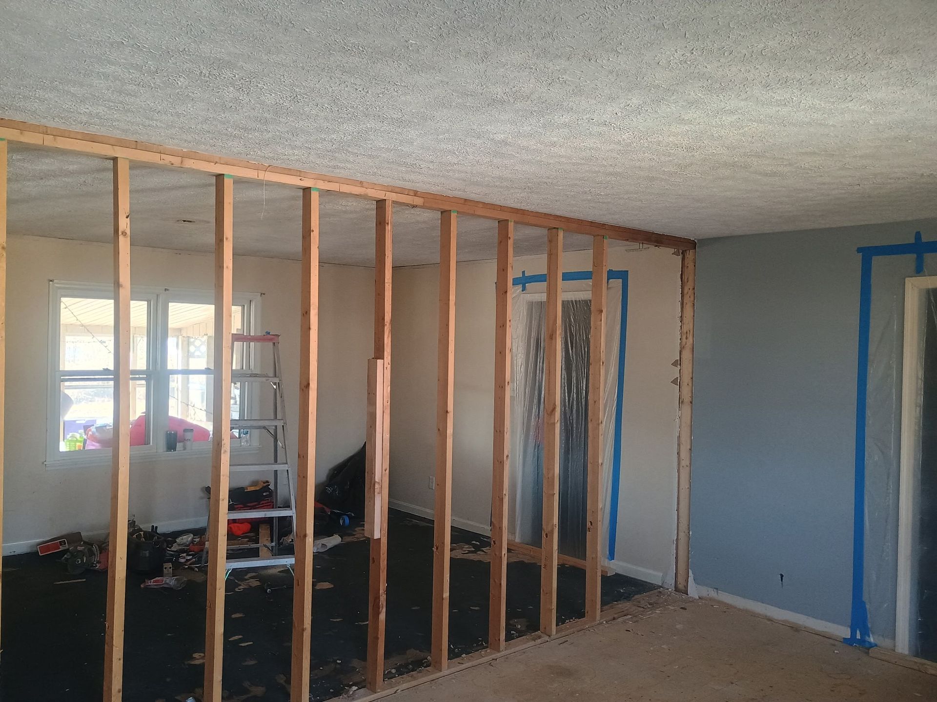 Interior under construction with exposed wood framing dividing a room, blue painter’s tape on walls and windows.