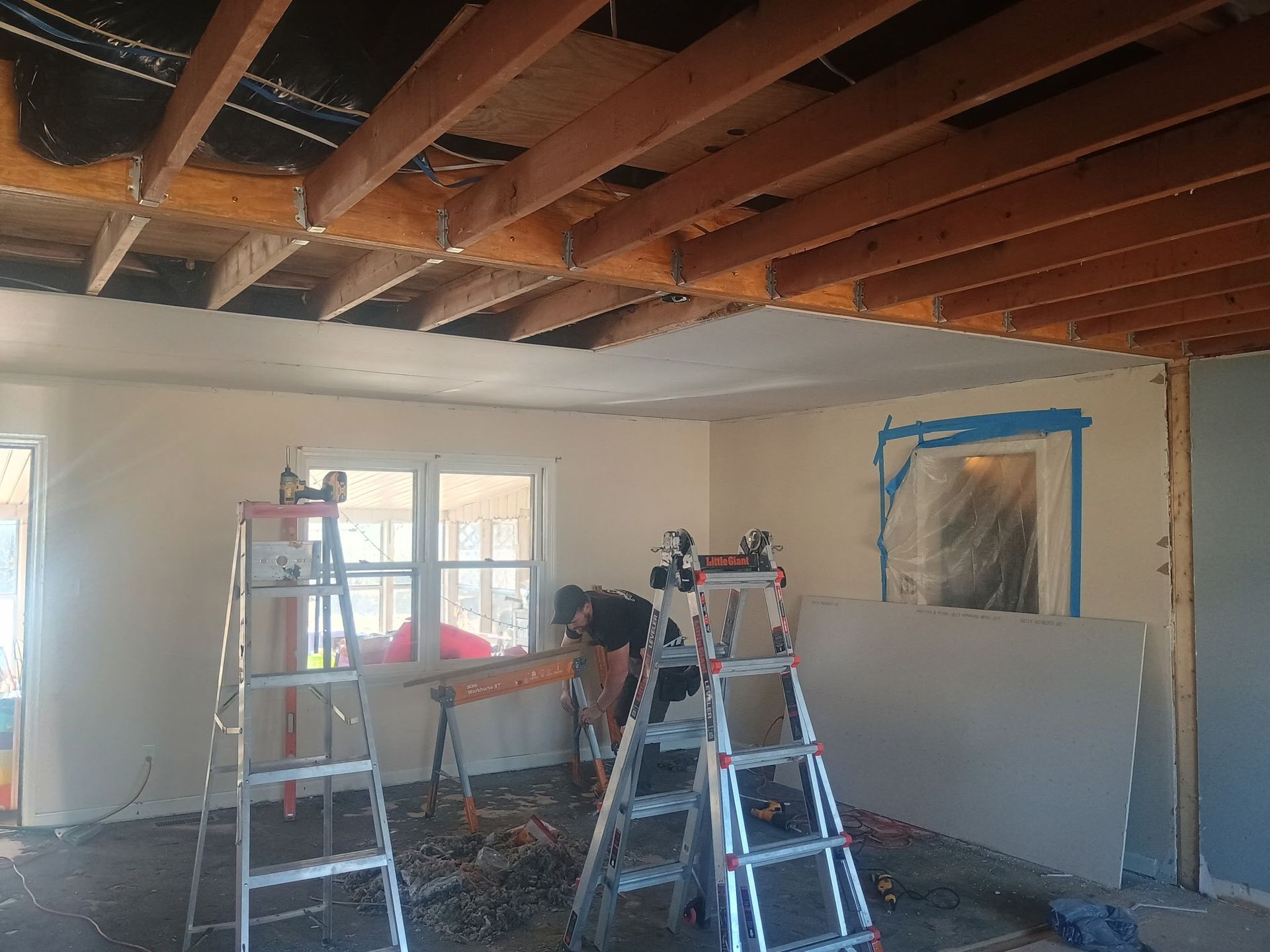 Room under renovation with exposed ceiling beams, ladders, and plastic-covered window and wall opening