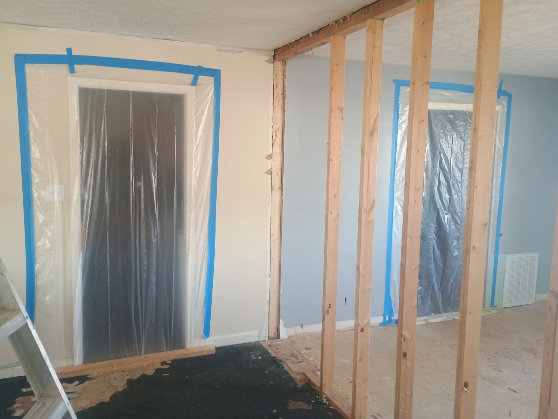 Interior room under renovation with blue-taped plastic-covered windows and exposed wooden framing.