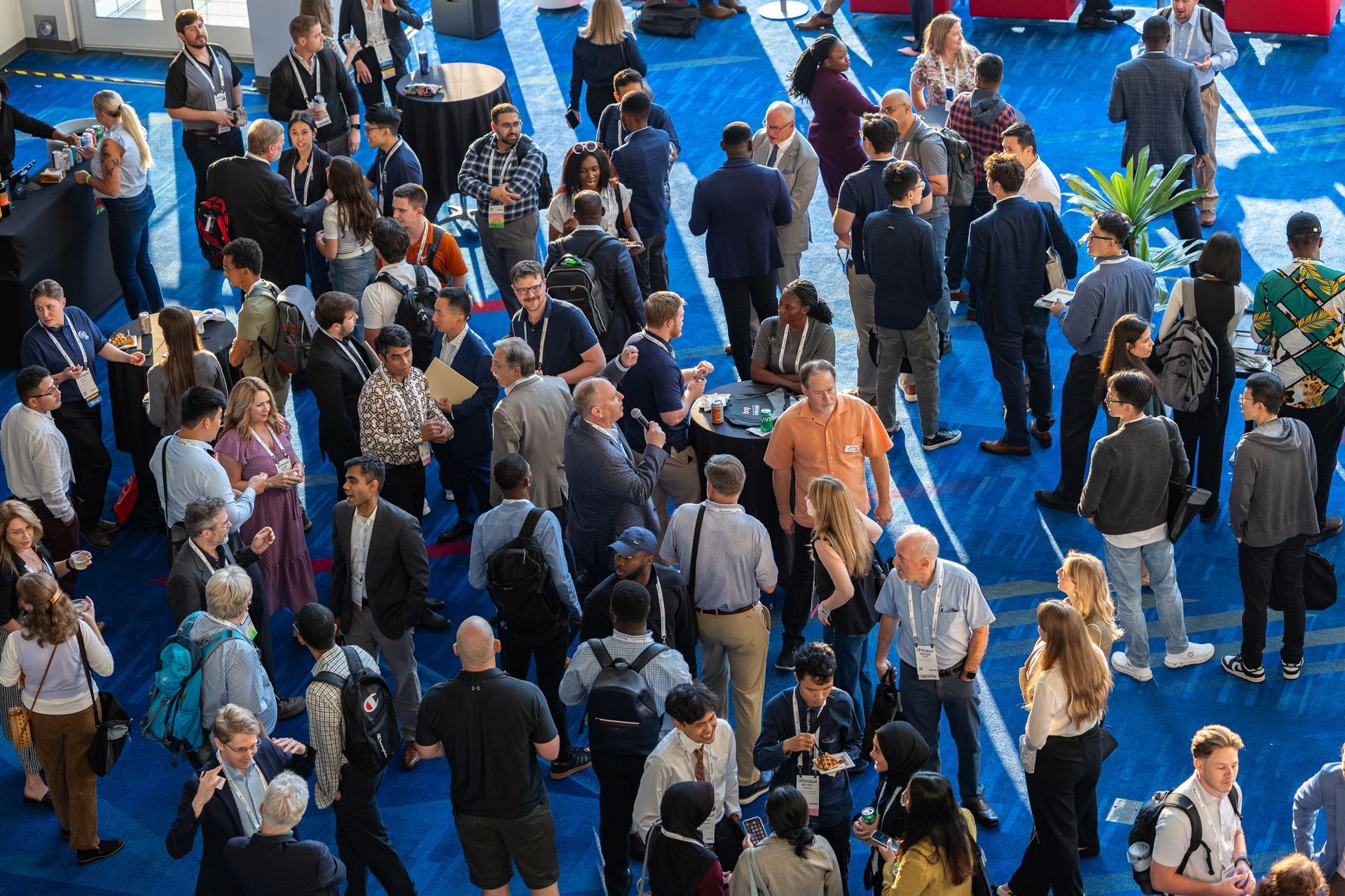 Large crowd of people at a conference or event on a blue carpeted floor, conversing and mingling.