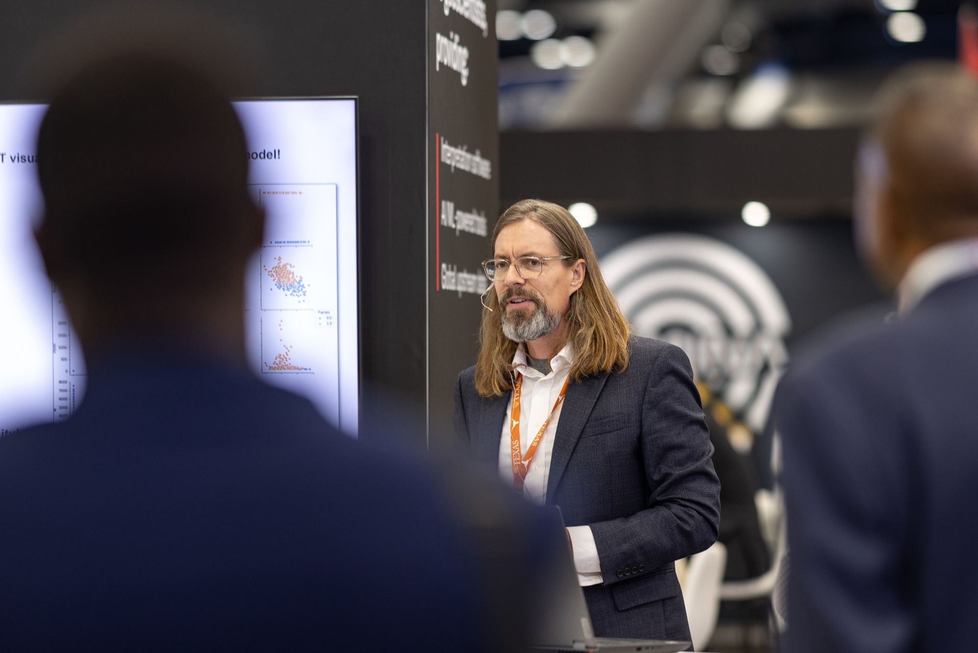 Man with beard presenting at a conference, pointing to a graph on a screen while two attendees listen.