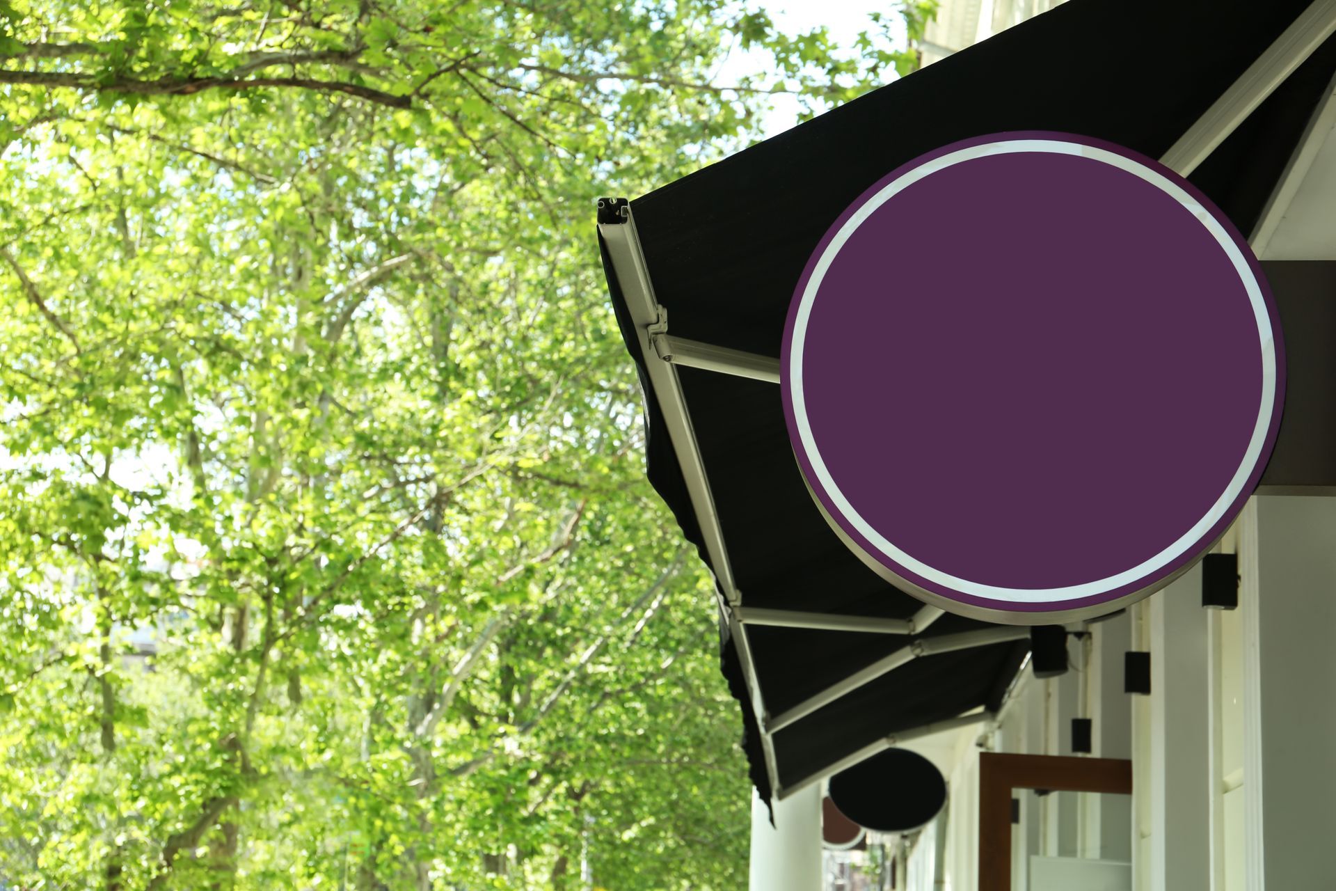A blank purple circular sign hangs from a black awning above a building storefront next to lush green trees.