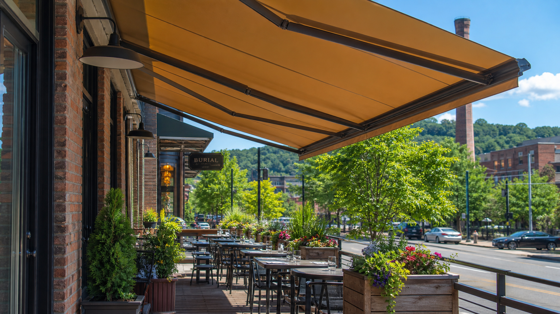 Outdoor café patio with tables under a tan awning, beside a sunny street and green trees
