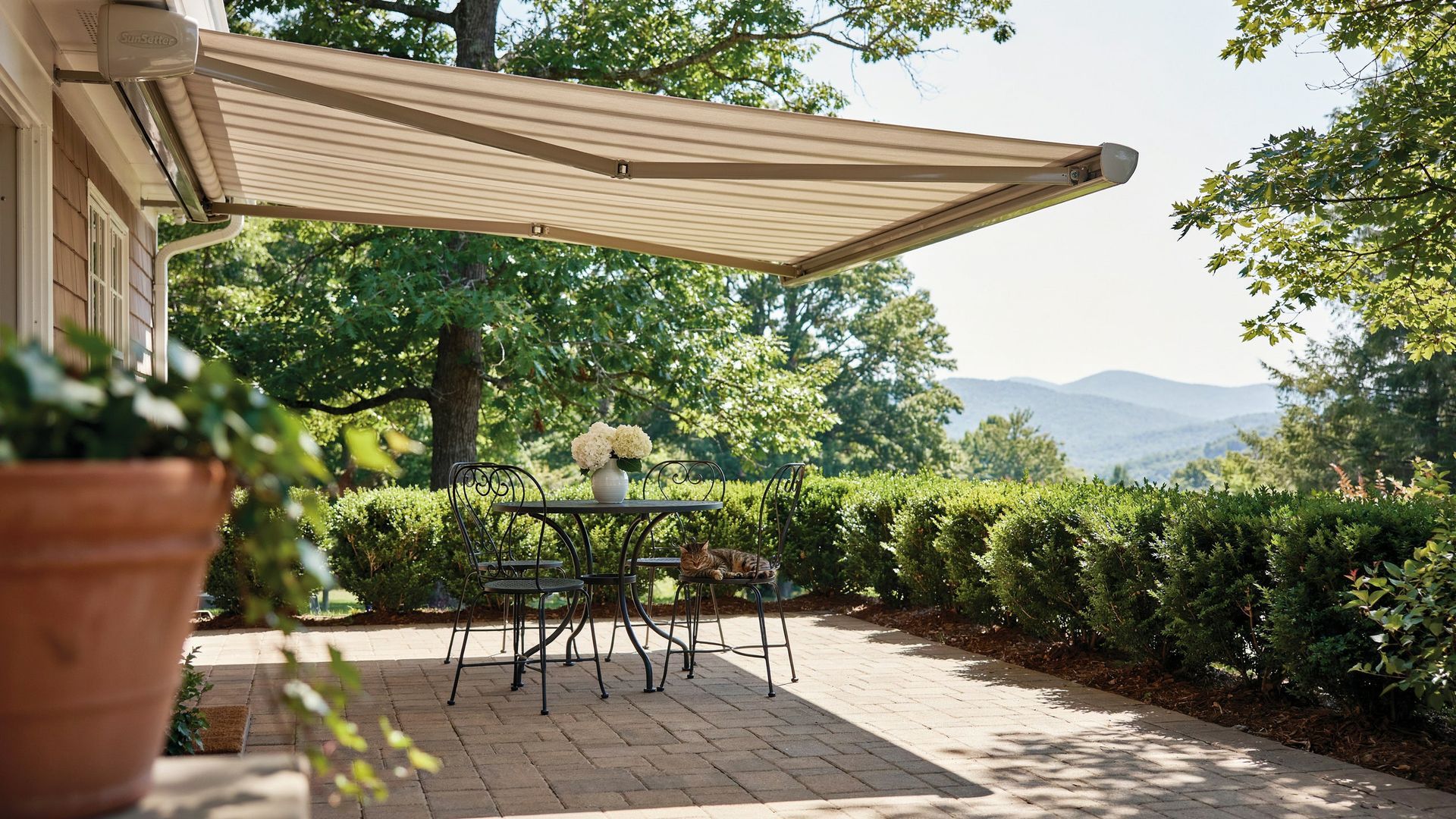 Shaded patio with a table and chairs beside a garden, under a striped awning on a sunny day