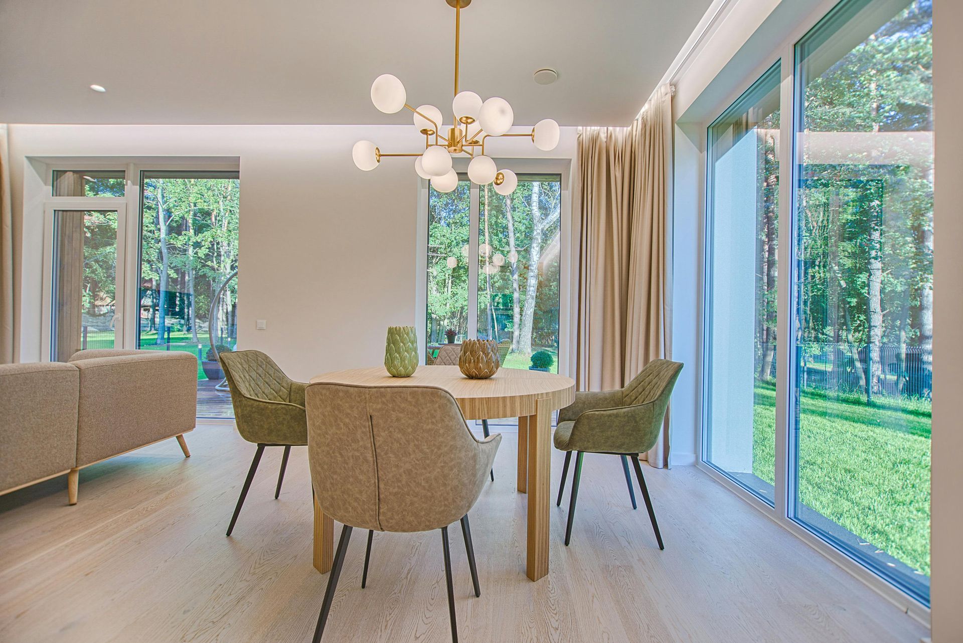 Dining room with round table, chairs, chandelier, and large windows overlooking greenery.