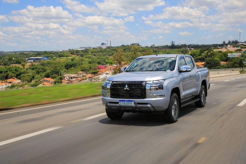 Caminhonete Mitsubishi prateada trafegando em uma estrada, com uma paisagem ao fundo sob um céu ensolarado.
